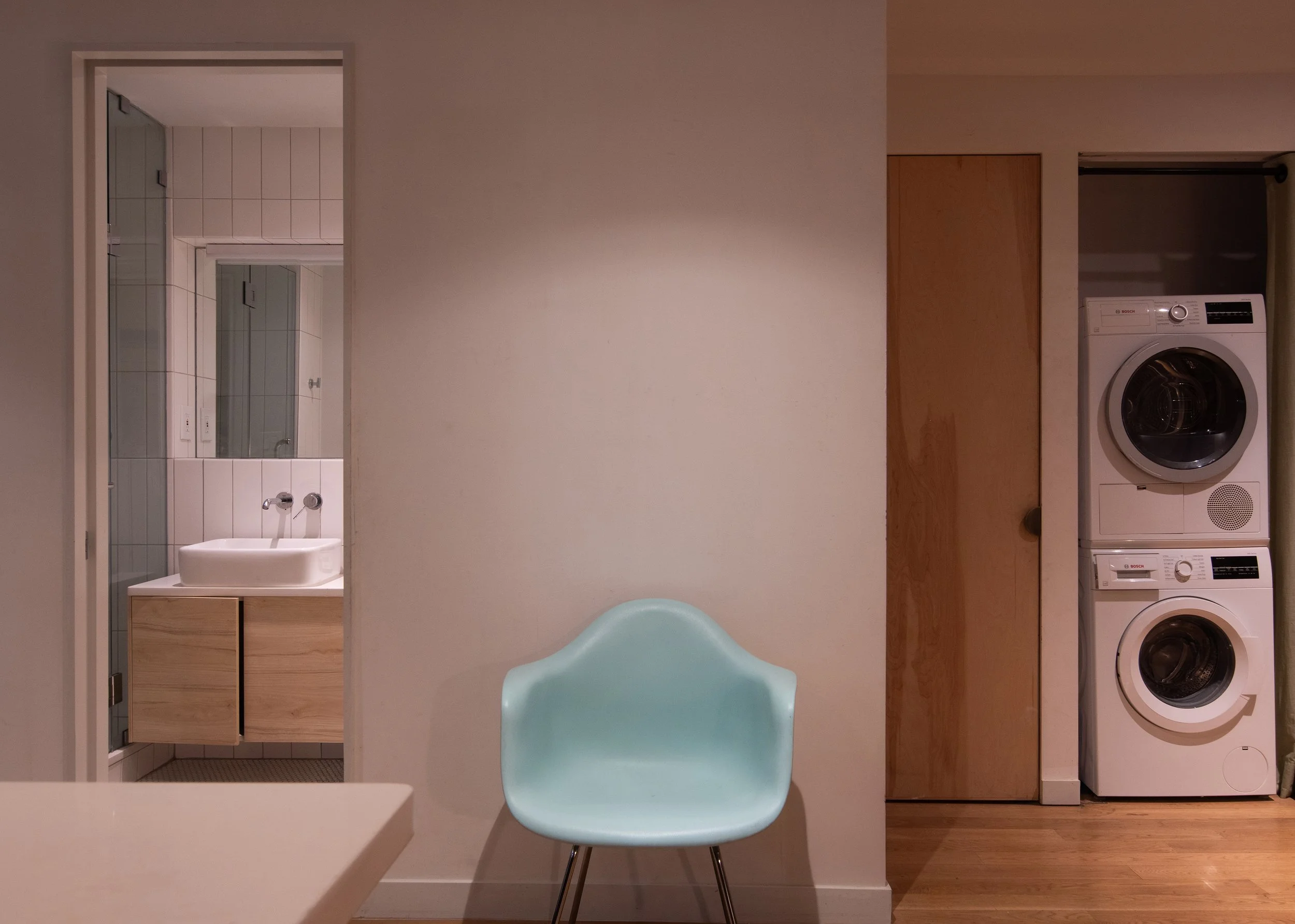 A light blue chair against a beige wall, with a doorway to a bathroom on the left and a stacked washer and dryer on the right.
