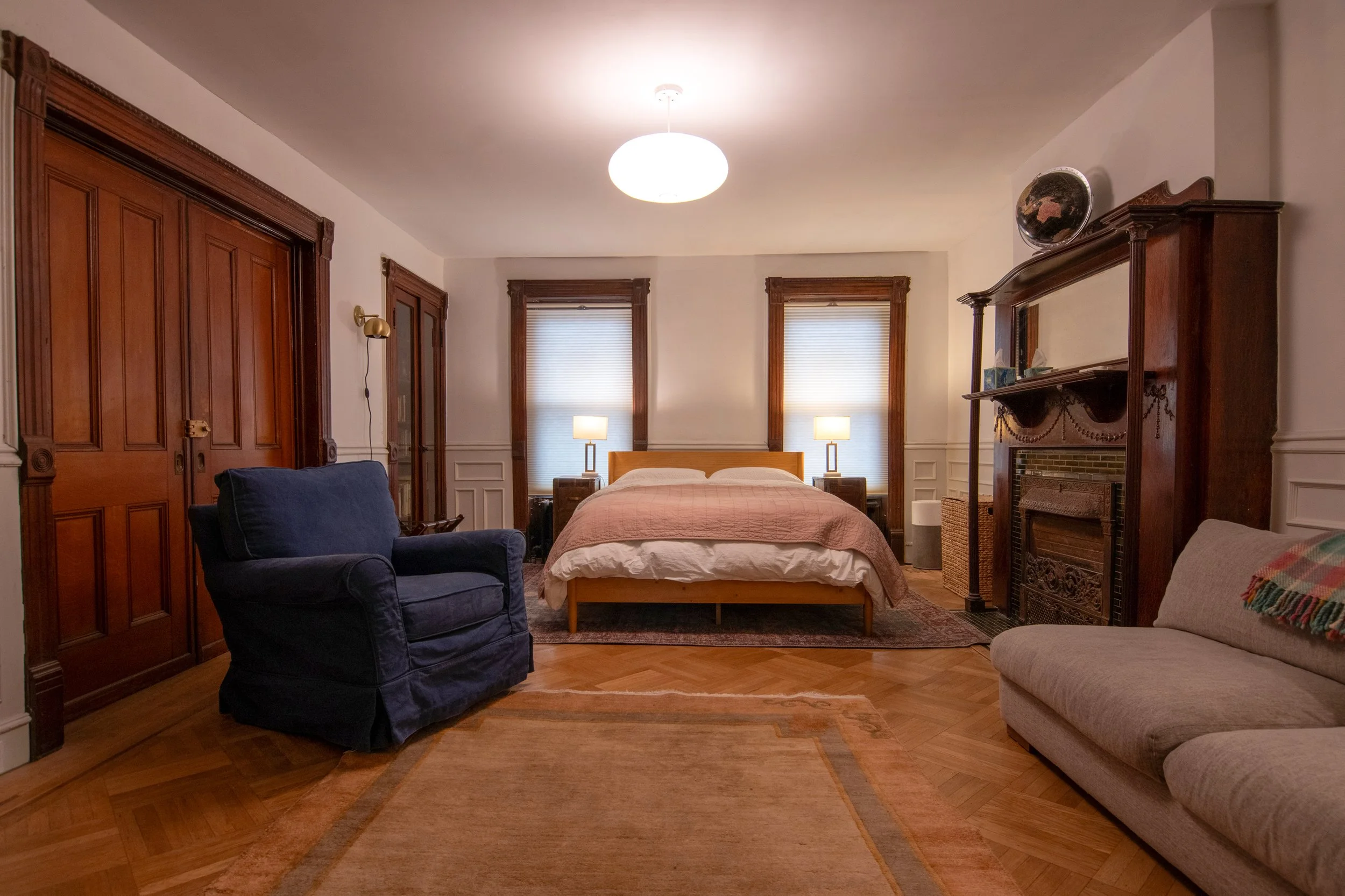 A cozy bedroom with a wooden bed, two nightstands with lamps, a fireplace with a wooden mantle, a navy armchair, a beige couch, and three windows with blinds.