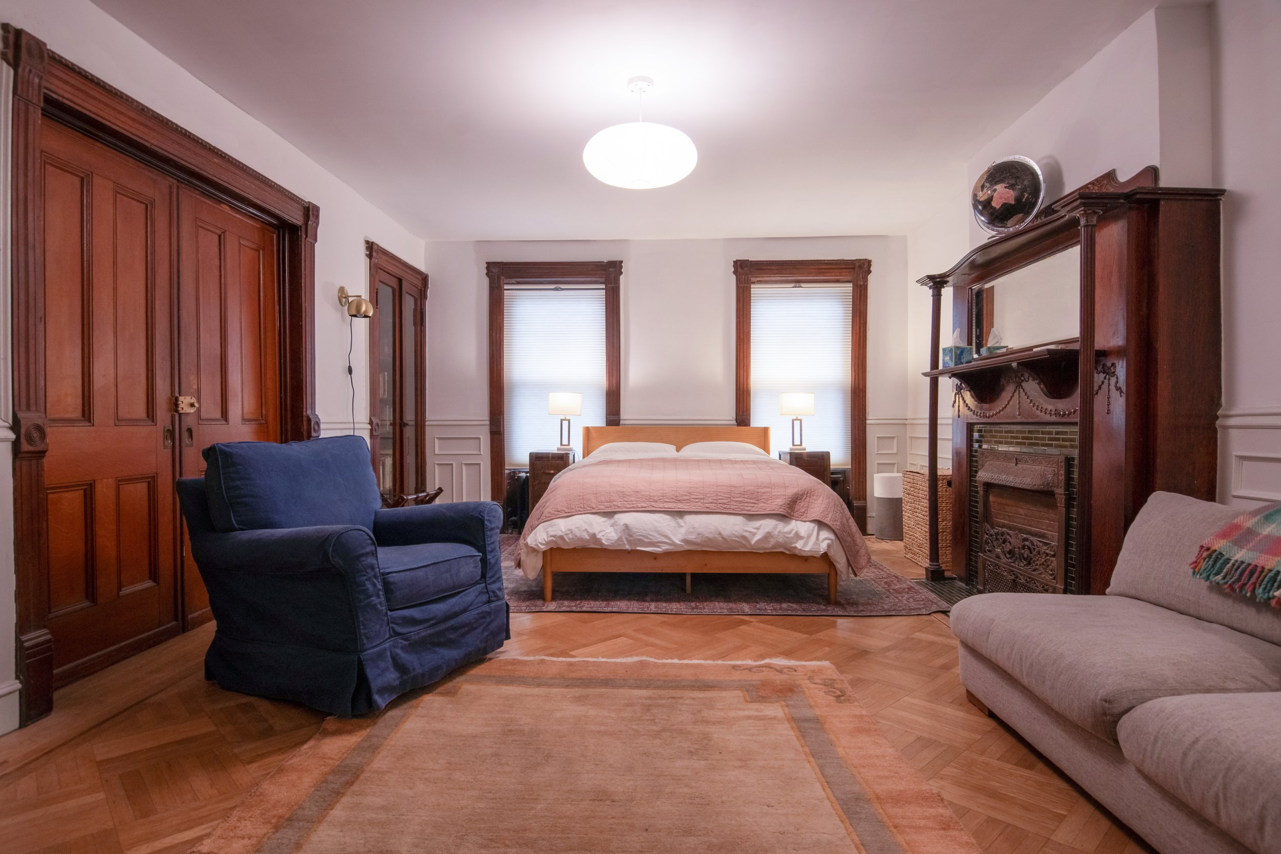 A cozy bedroom with a wooden bed, two nightstands with lamps, a fireplace with a wooden mantle, a navy armchair, a beige couch, and three windows with blinds.