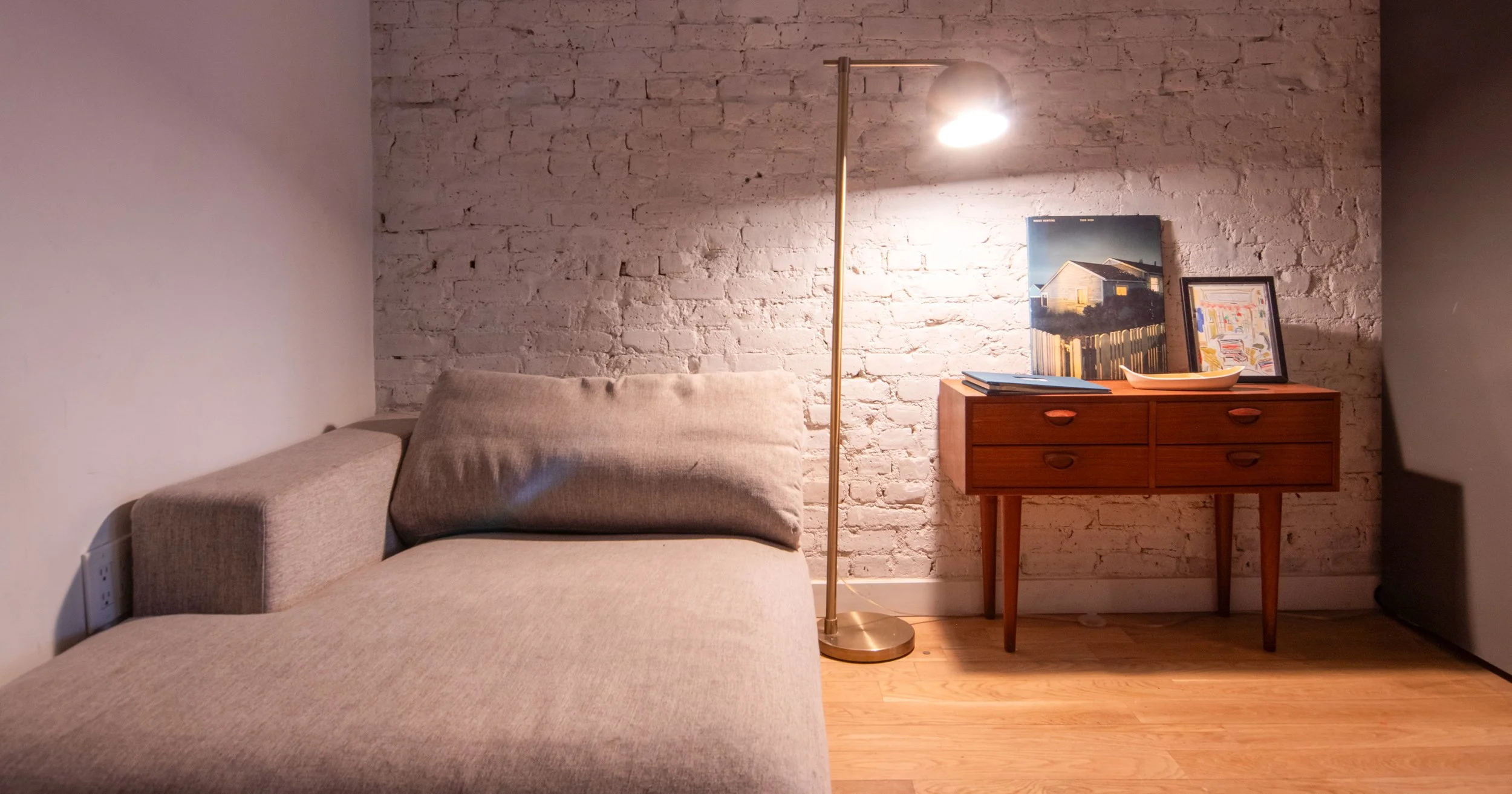 Living room corner with beige L-shaped sofa, a brass floor lamp, a wooden sideboard with framed artwork, a large framed picture, and decorative plates, against a white brick wall and wooden floor.