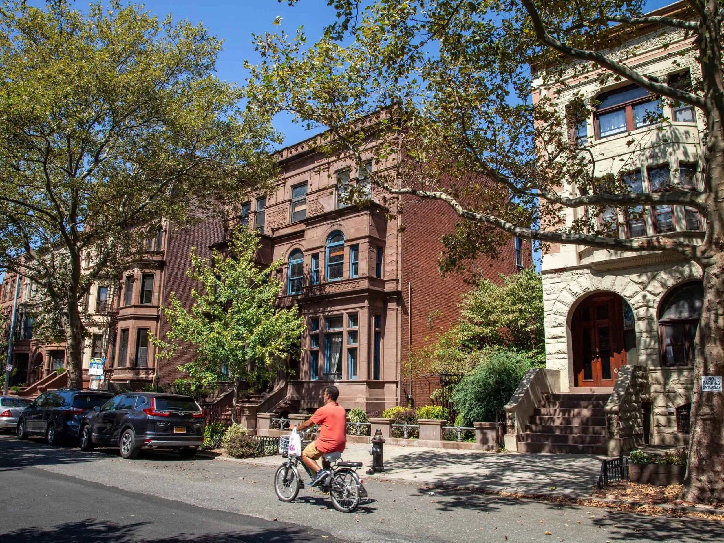 A city street with old brownstone-style houses, trees, parked cars, and a person riding a bicycle on the sidewalk.