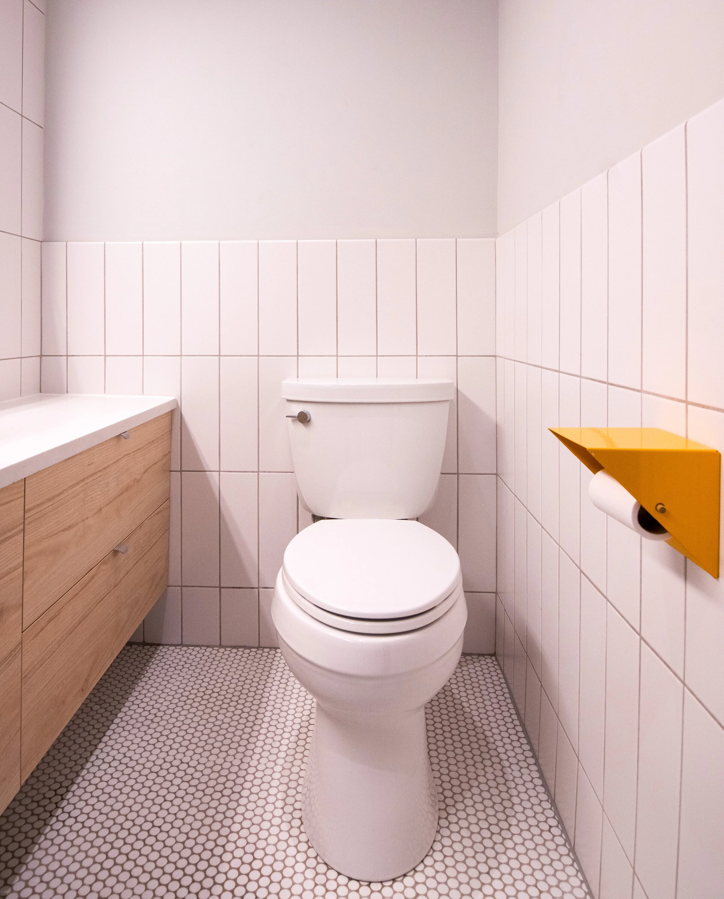 A small bathroom with a white toilet, light-colored wooden cabinet on the left, white tiled walls, and a yellow toilet paper holder on the right wall.