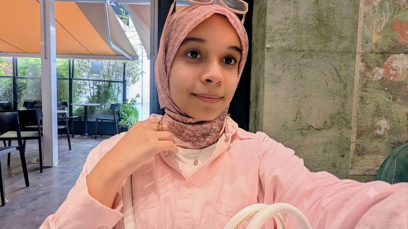A young girl with a light skin tone wearing a patterned black jacket, a white and pinkish hijab, sunglasses on her head, and a blue lanyard around her neck taking a selfie in a park with manicured flower beds, large trees, and a building with arches in the background.