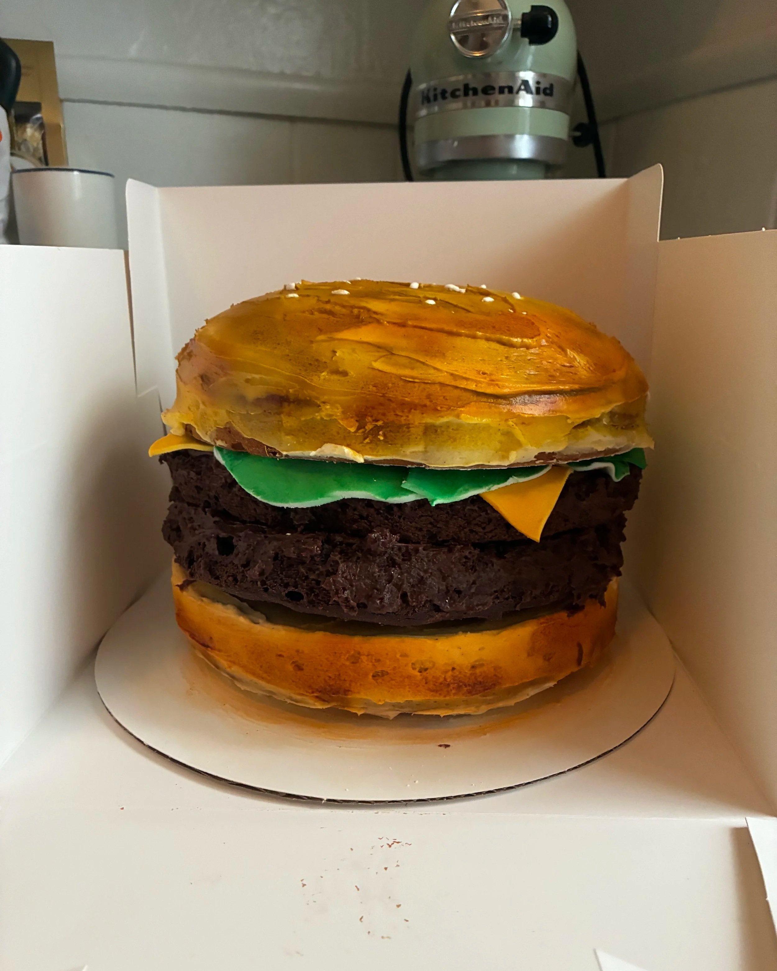 A burger-shaped cake with layers resembling a burger, including a bun, green and yellow fondant toppings, a chocolate layer representing the patty, and additional cake layers, in a white box with a KitchenAid mixer in the background.