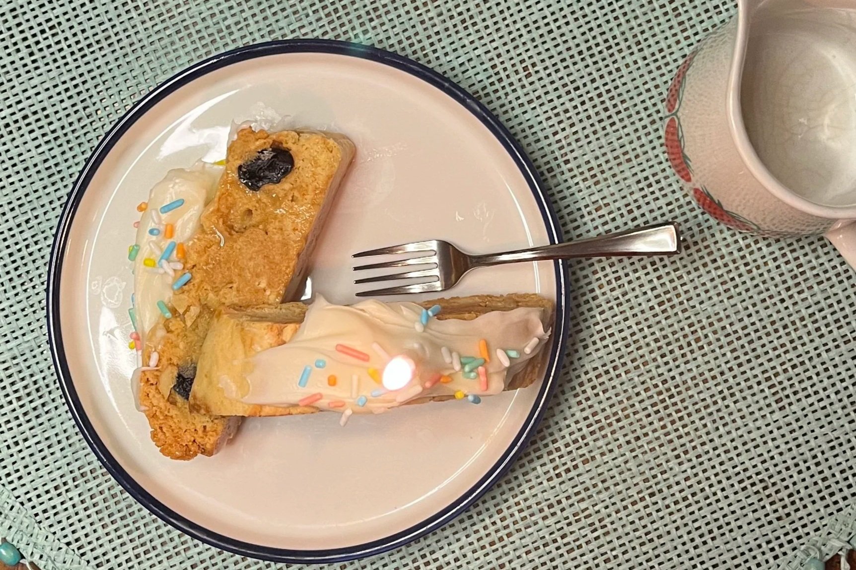 A plate with two slices of birthday cake with sprinkles, one slice with cream and fruit toppings, a fork, and a mug of water on a green tablecloth.