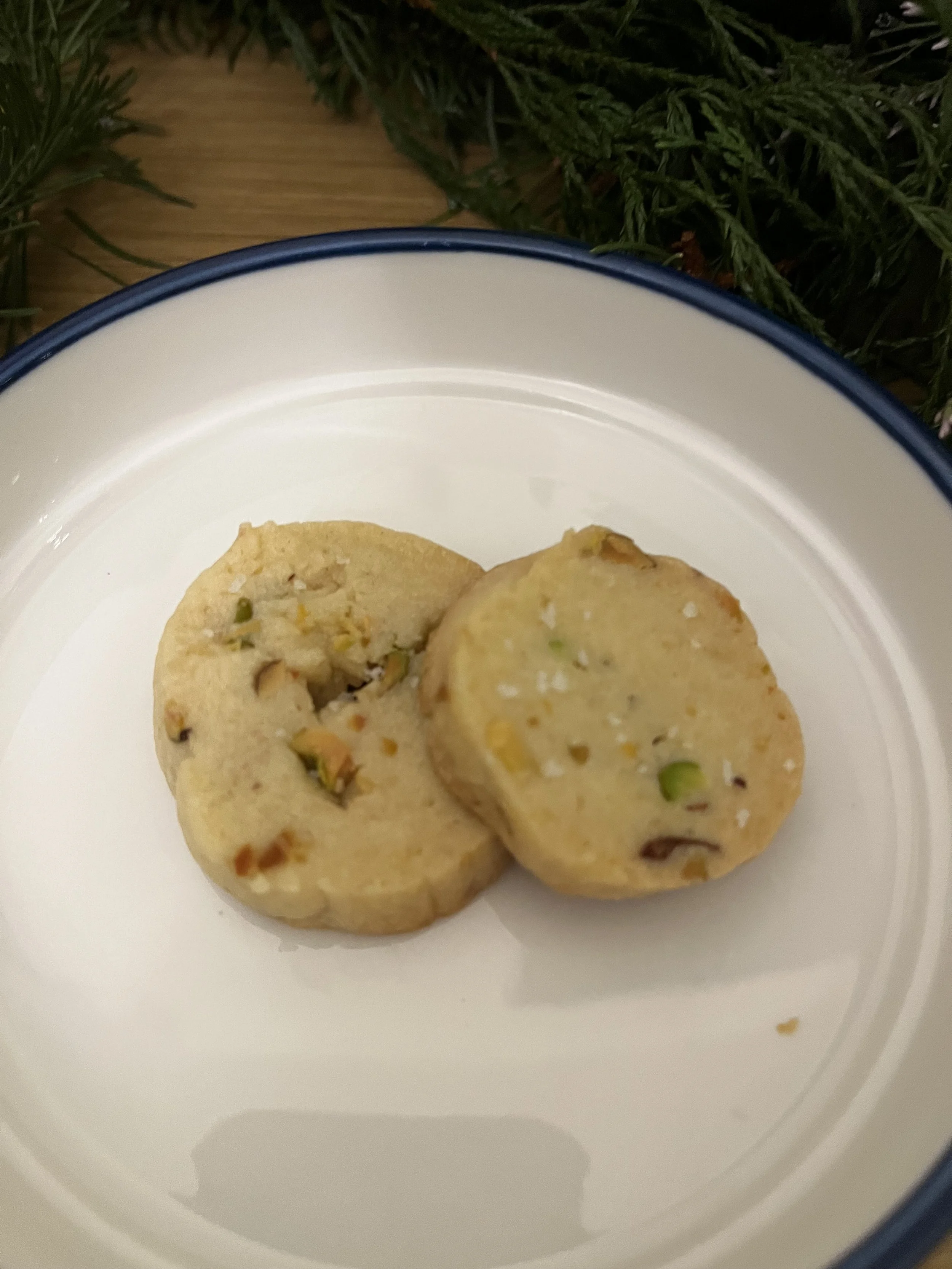 Two pistachio-flavored cookies on a white plate with a blue rim, with greenery in the background.