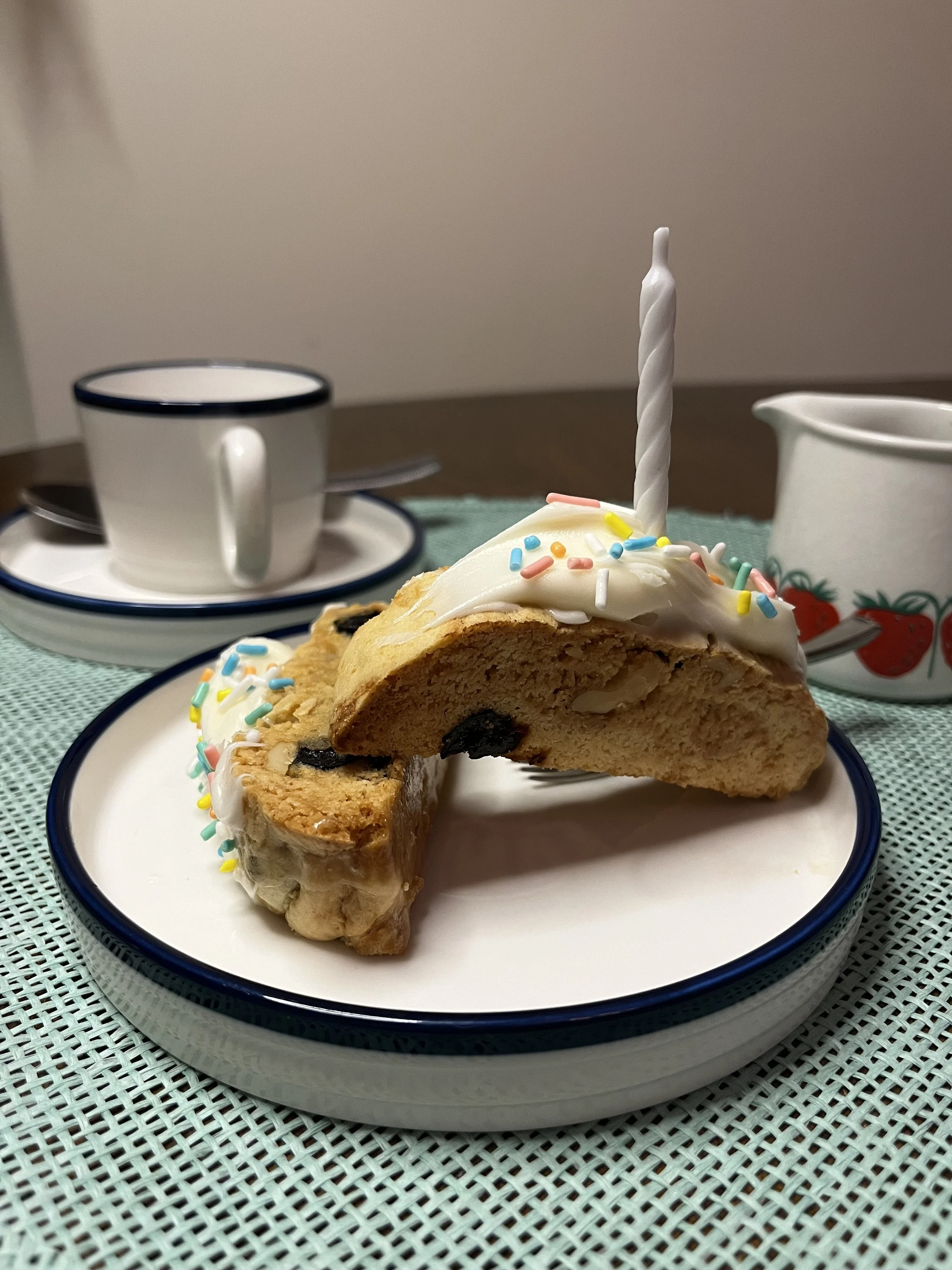 A slice of birthday cake with white frosting, colorful sprinkles, and a lit birthday candle on top, placed on a white plate with a black rim. In the background, there are a white cup with a saucer and a small pitcher with a strawberry pattern, all on