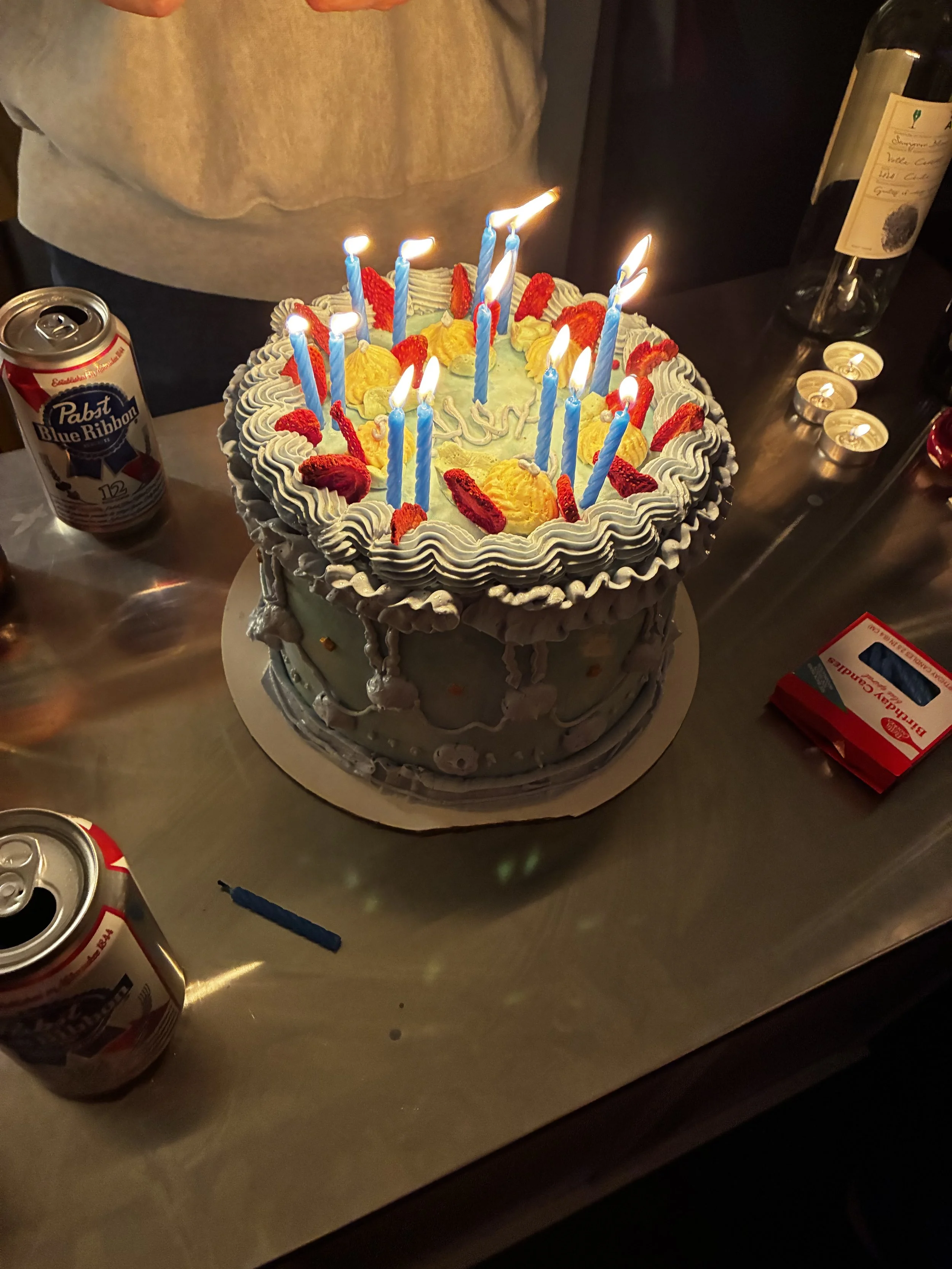 A birthday cake with lit candles, decorated with strawberries and yellow cream topping, placed on a table with drinks and small candles around it.