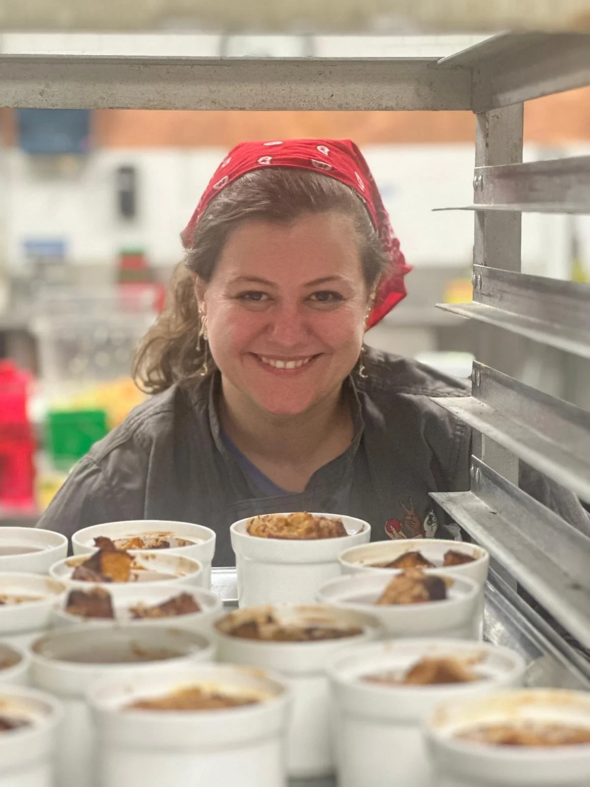 A woman smiling behind a row of white cups filled with food, wearing a red bandana and a black shirt, in a kitchen or food preparation area.