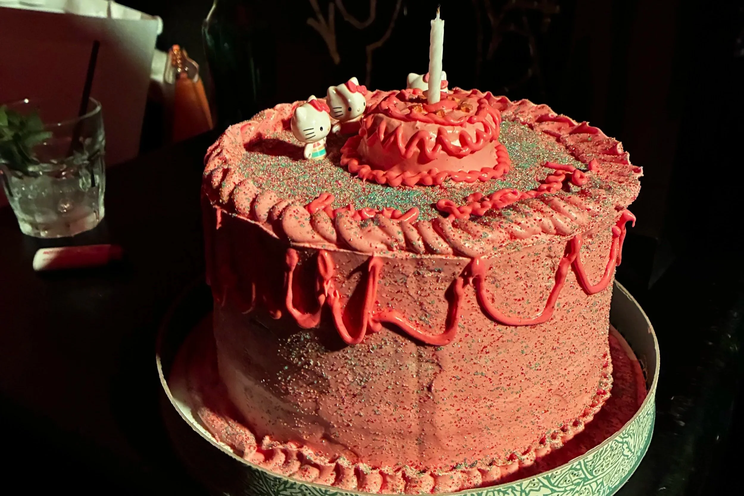 Pink birthday cake decorated with glitter, pink icing, Hello Kitty figures, and a candle on top.