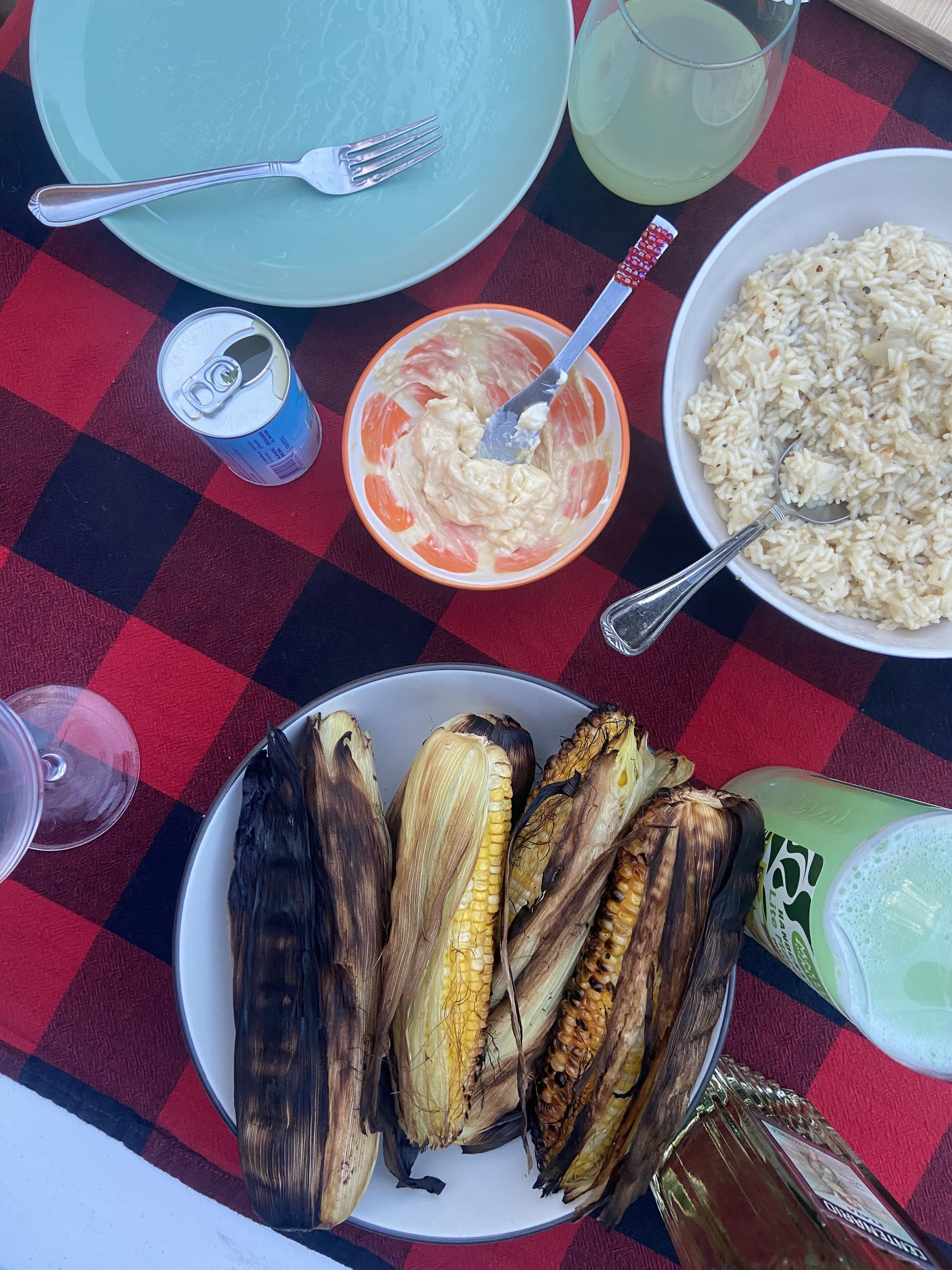 A table set outdoor with grilled corn on the cob, rice, potato salad, and drinks, on a red and black checkered tablecloth.