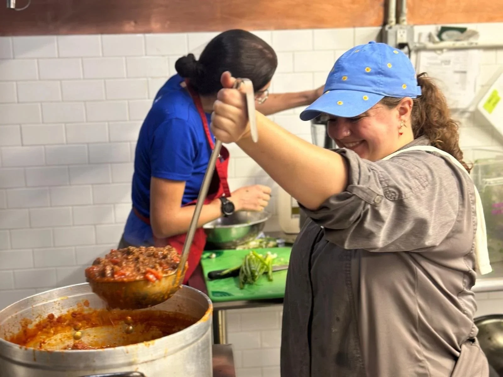 A woman wearing a blue cap and gray shirt smiling as she adds ingredients to a pot in a kitchen with a woman in the background cooking.