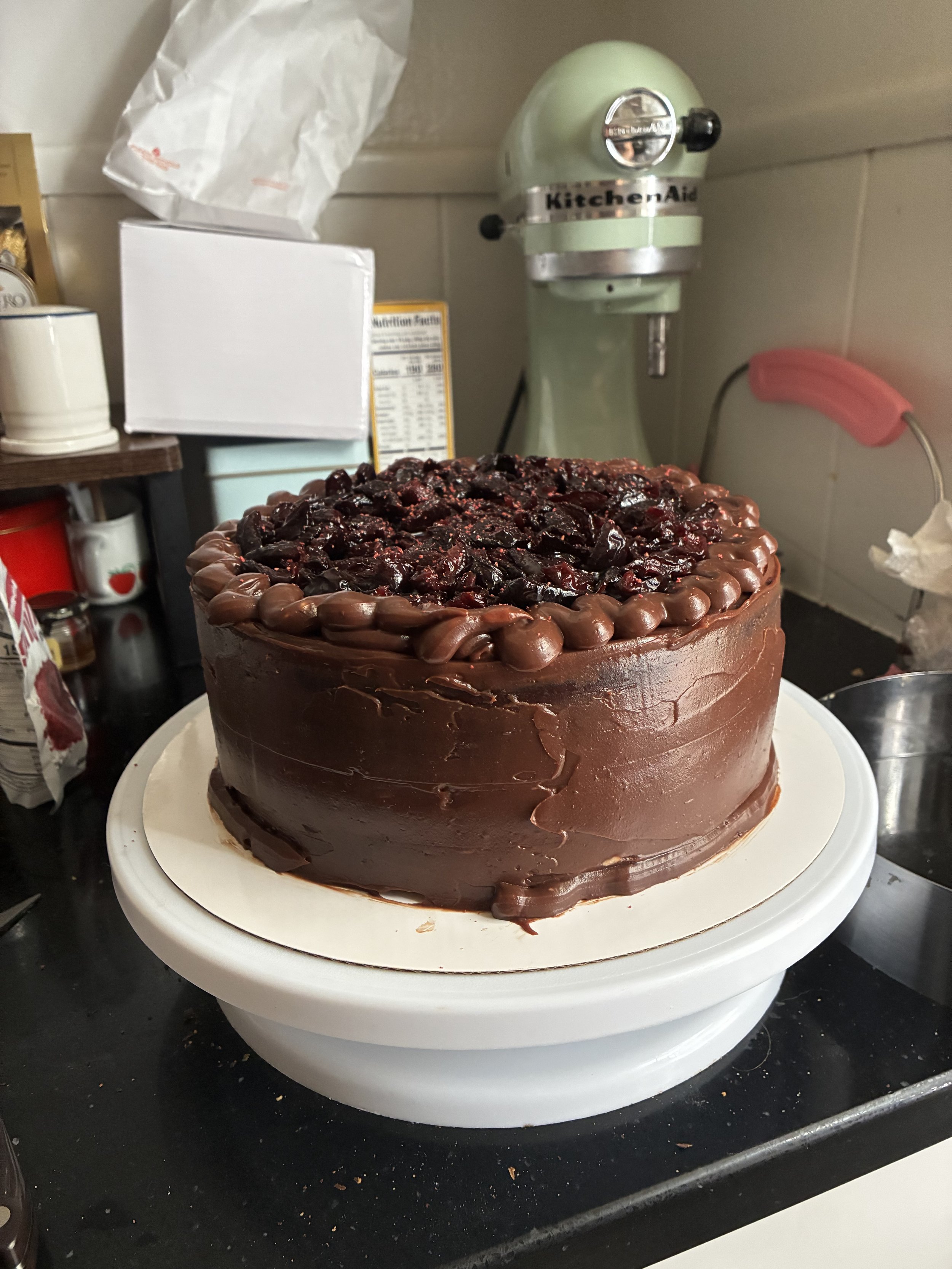 A large chocolate cake with a glossy chocolate frosting, topped with cherries and a layer of cherry filling, sits on a cake turntable in a kitchen.