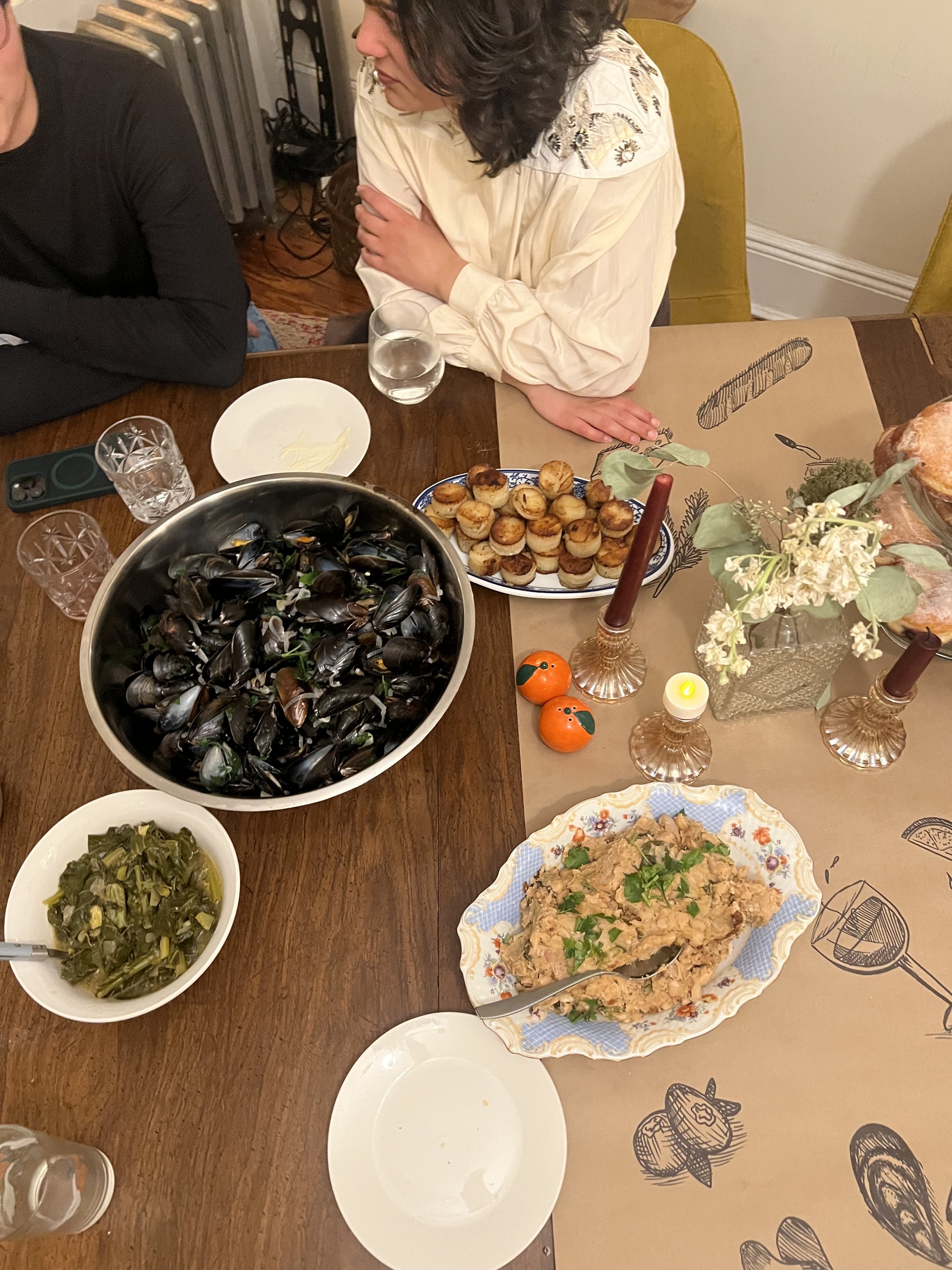 A dinner table with a bowl of cooked mussels, a plate of small stuffed mushrooms, a bowl of cooked greens, and a dish of tuna salad. There are glasses of water, decorative candles, and small orange-shaped salt and pepper shakers.