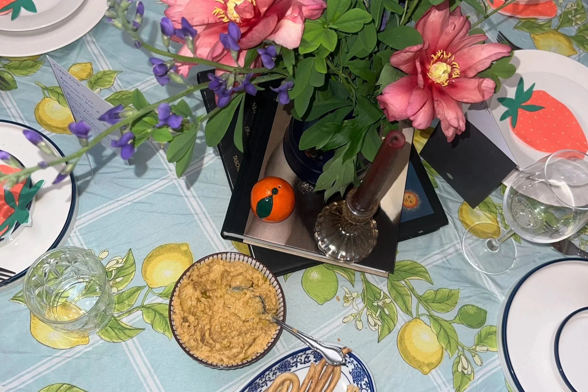 A table set with plates, glasses, and a floral centerpiece. The centerpiece includes pink flowers and purple sprigs in a glass vase, with candles and small decorative items nearby. There is also a bowl of dip or spread and some breadsticks on the table.