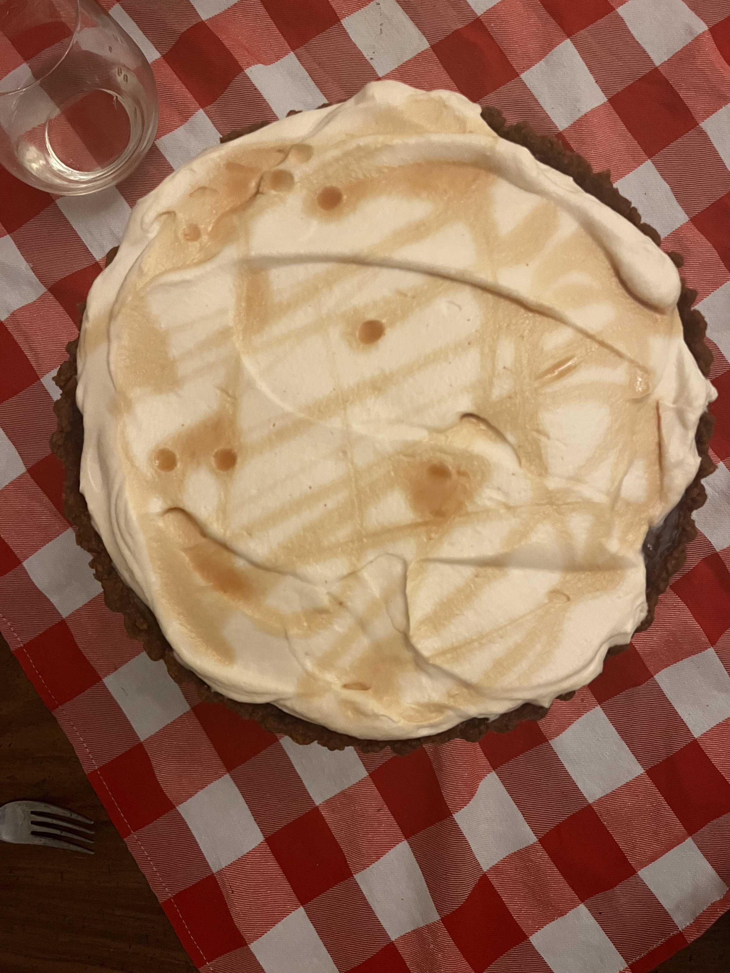 A pie with a graham cracker crust and a creamy filling topped with caramel sauce, on a checkered red and white tablecloth.