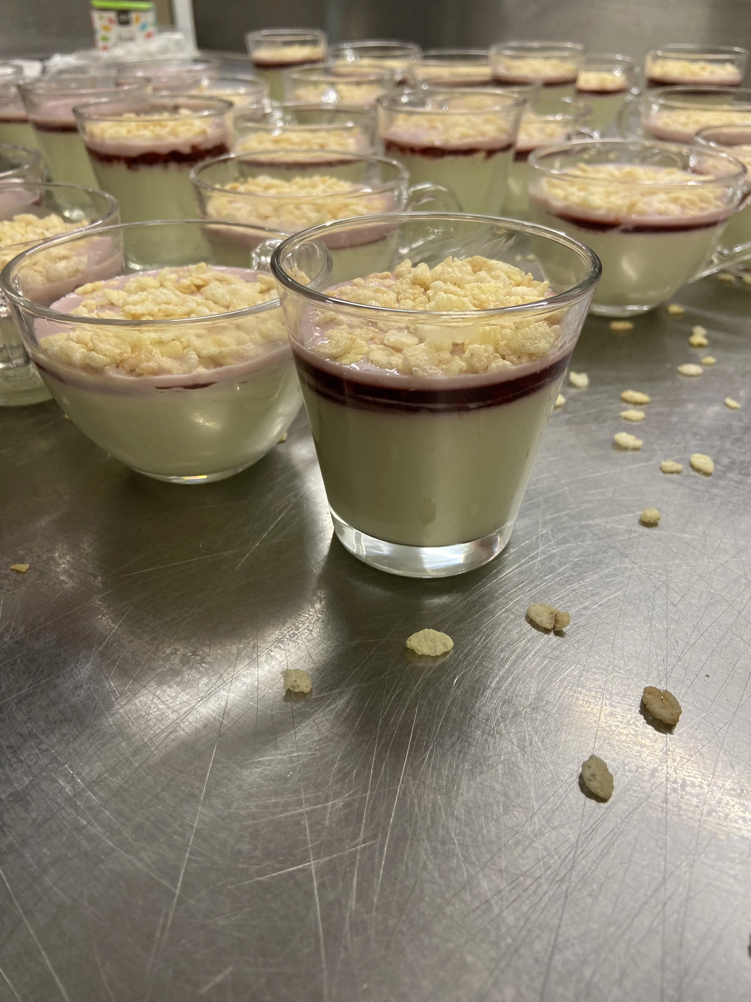 Several glass cups filled with layered dessert with a yellow custard or pudding base, topped with a layer of red berry sauce, and sprinkled with crumbled white topping, possibly white chocolate or meringue.