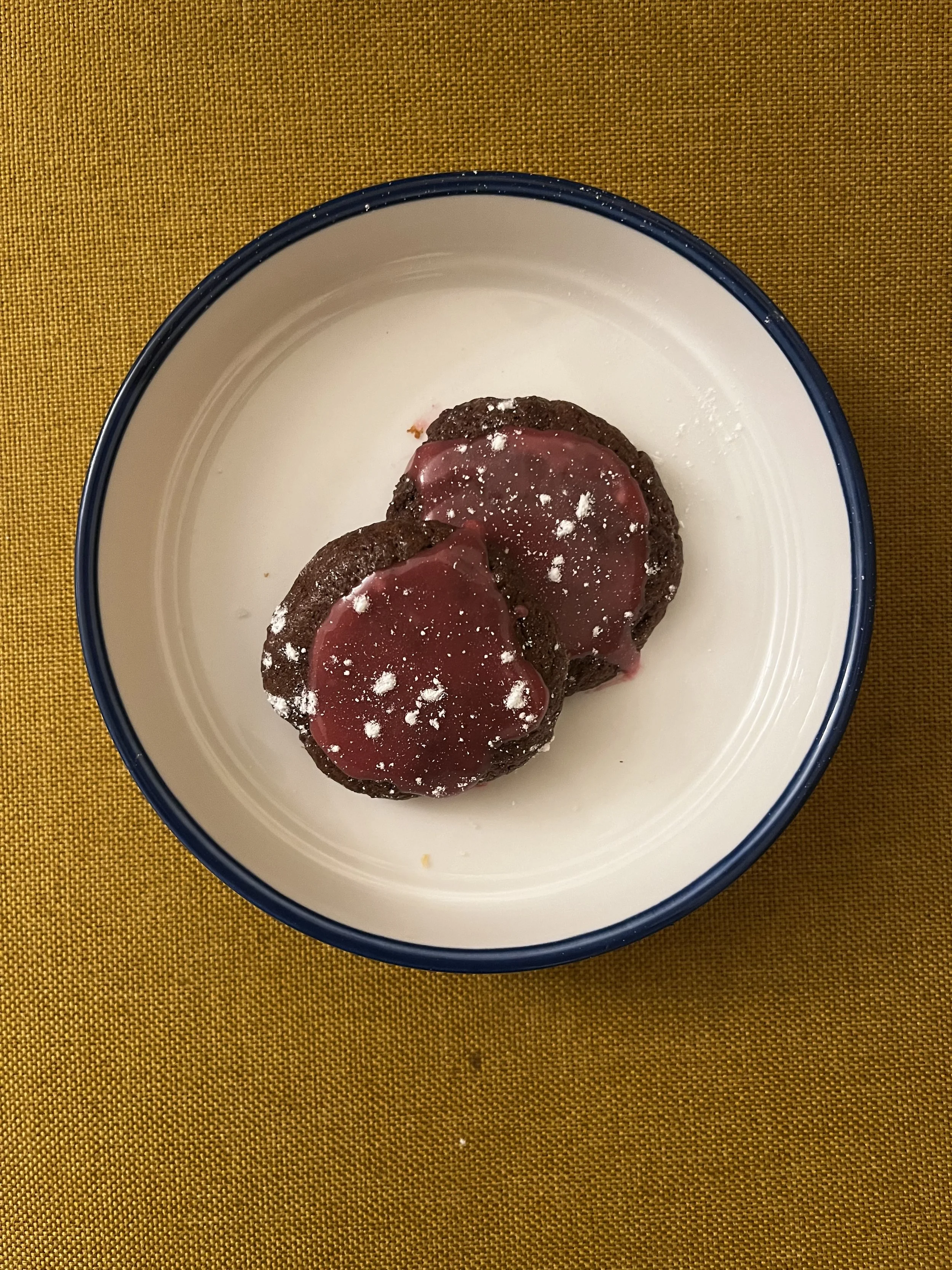 Two cookies topped with red jelly and sprinkled with powdered sugar on a white plate with a blue rim, placed on a yellow textured surface.