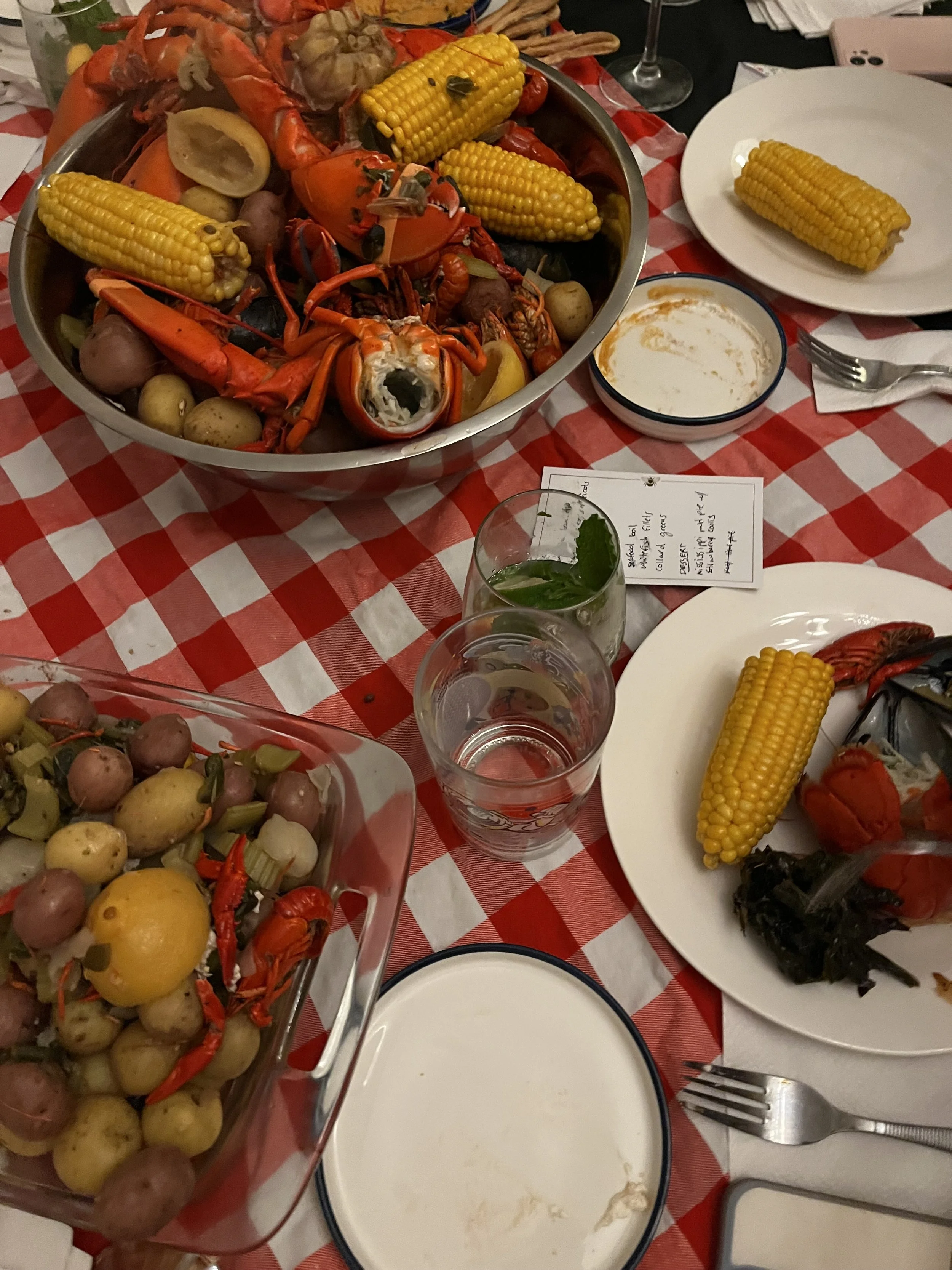 Seafood boil with crab, crawfish, and corn on a red and white checkered tablecloth, with sides of potatoes, lemon, and vegetables, along with glasses of water and summer drinks.