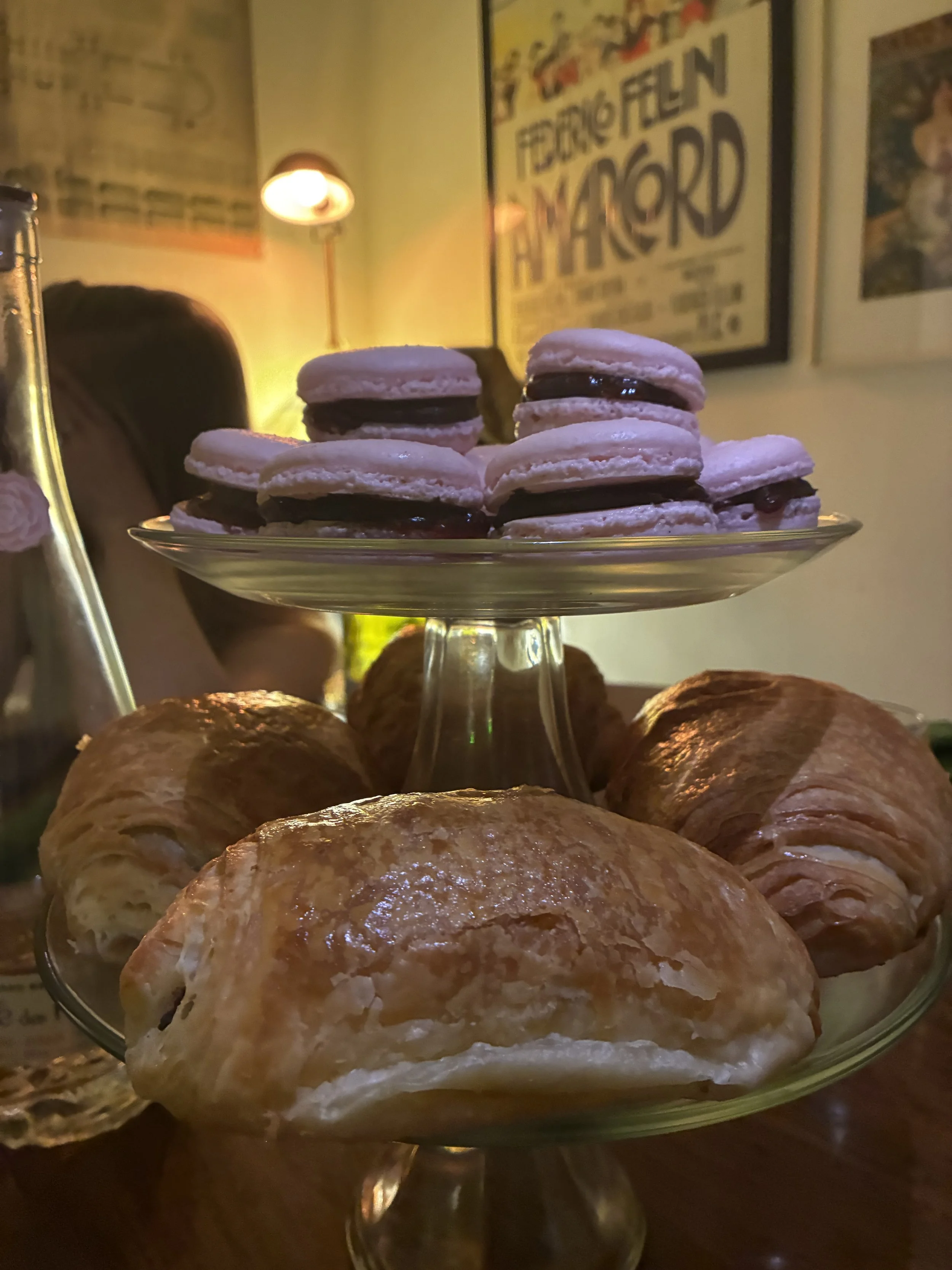 Assorted pastries including croissants and macarons on a glass stand in a cafe or restaurant setting.
