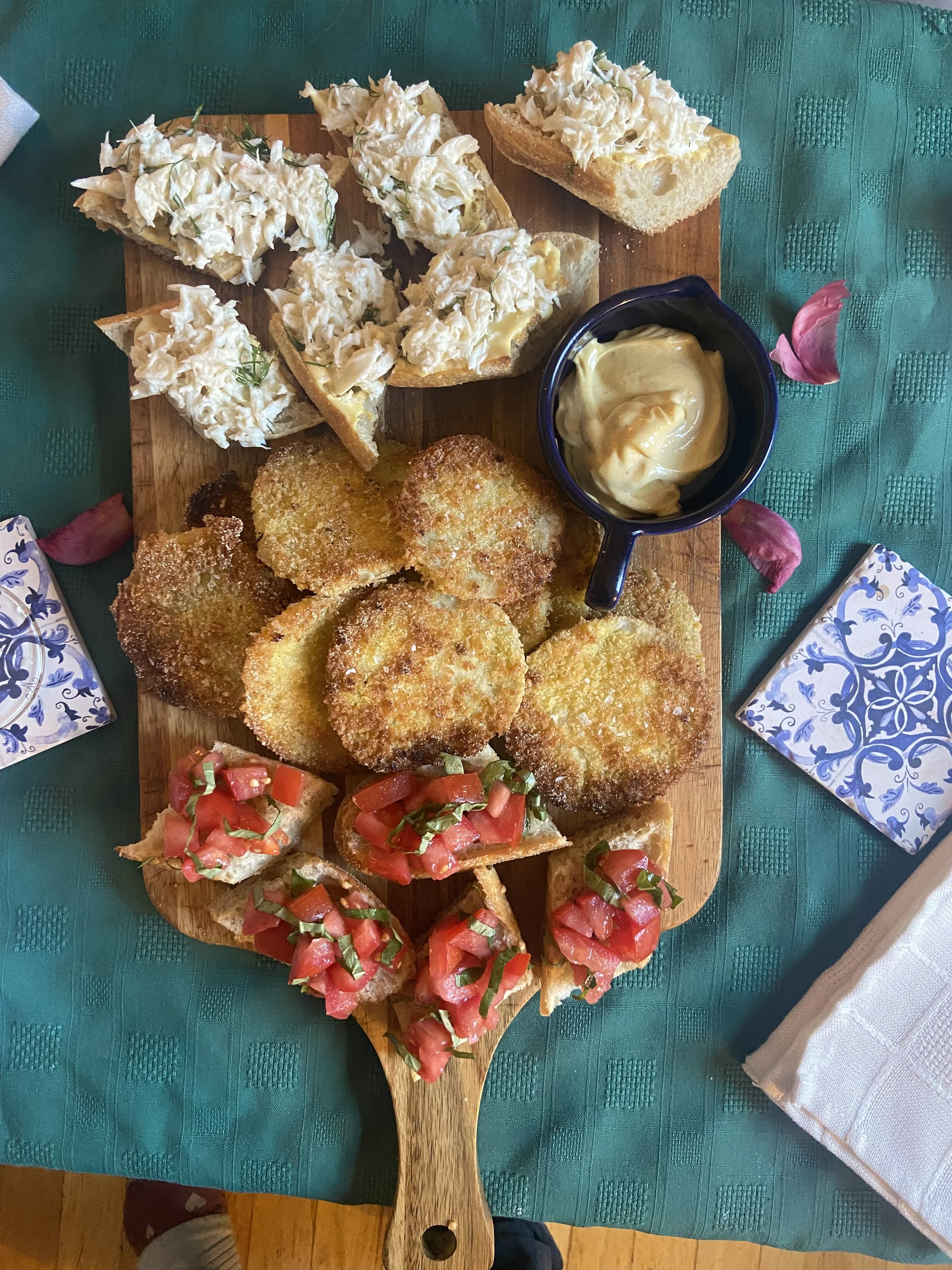 A wooden serving platter with toasted bread topped with crab salad, breaded fried cutlets, tomato bruschetta, and a small bowl of mayonnaise with a hellmann's label. The platter is on a green tablecloth with pink flower petals and blue patterned napk