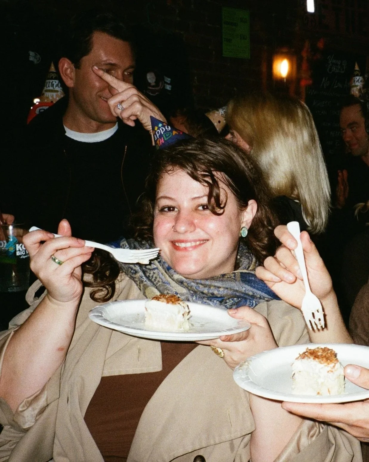 A woman with curly hair, wearing earrings and a beige coat, holds a plate with a slice of cake and a fork, smiling at the camera while at a celebration event with people in the background, some wearing birthday hats.