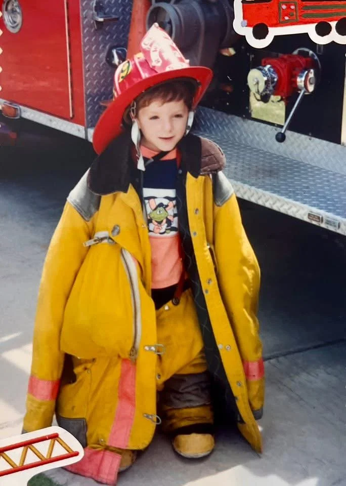 Michael as a young boy dressed as a firefighter, wearing a yellow firefighter jacket, helmet, and boots, kneeling in front of a fire truck.