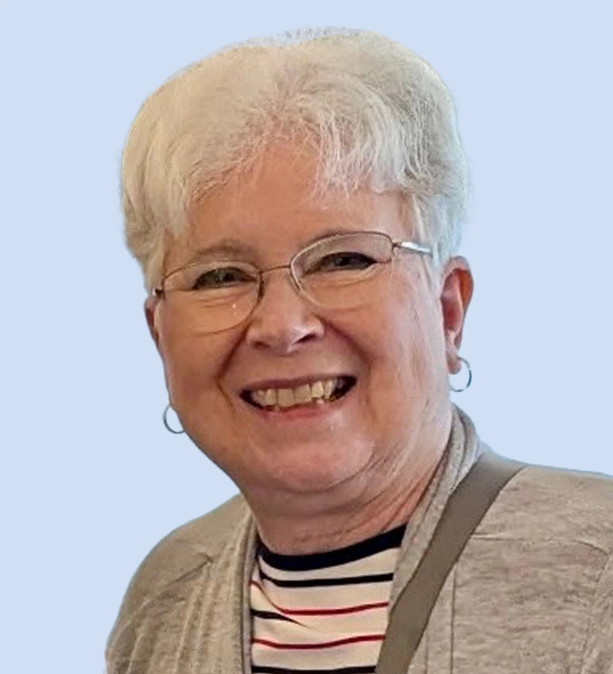 Board member Judi. Smiling elderly woman with short white hair, glasses, hoop earrings, wearing a striped shirt and beige cardigan, against a light blue background.
