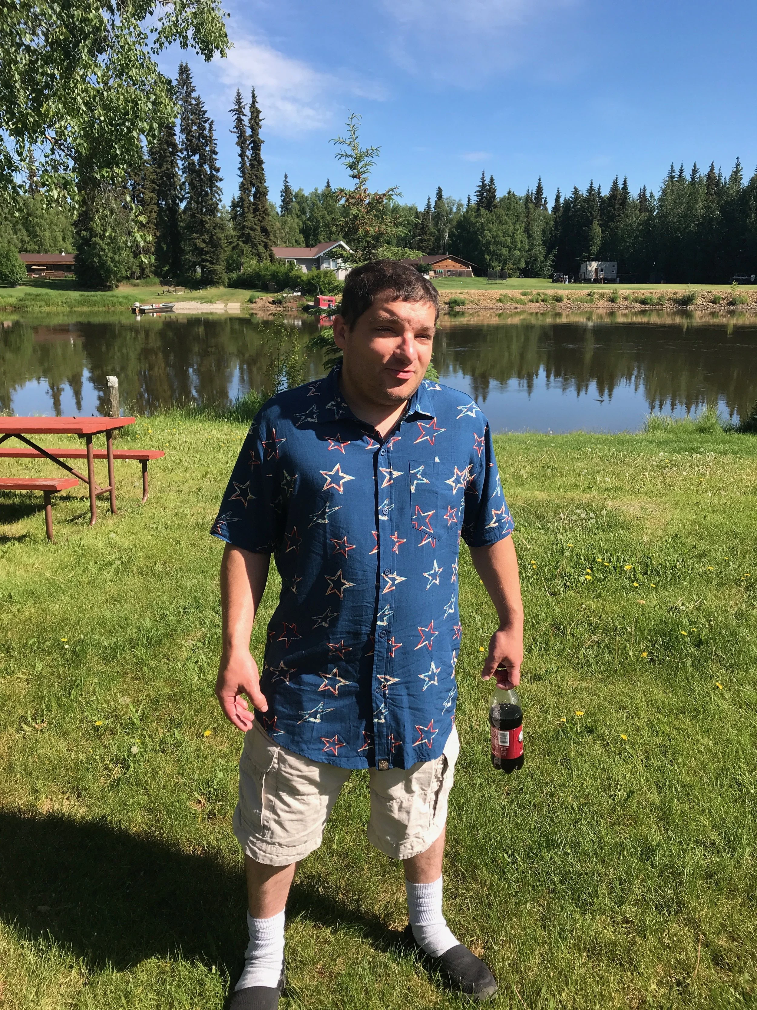 Michael standing outdoors next to a lake, holding a bottle of soda, wearing a blue shirt with star patterns, khaki shorts, white socks, and black shoes.