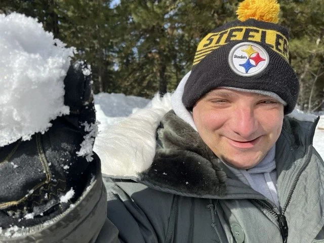 Michael outdoors in the snow, wearing a Pittsburgh Steelers beanie with a yellow pom-pom, smiling, with a snowball in his gloved hand