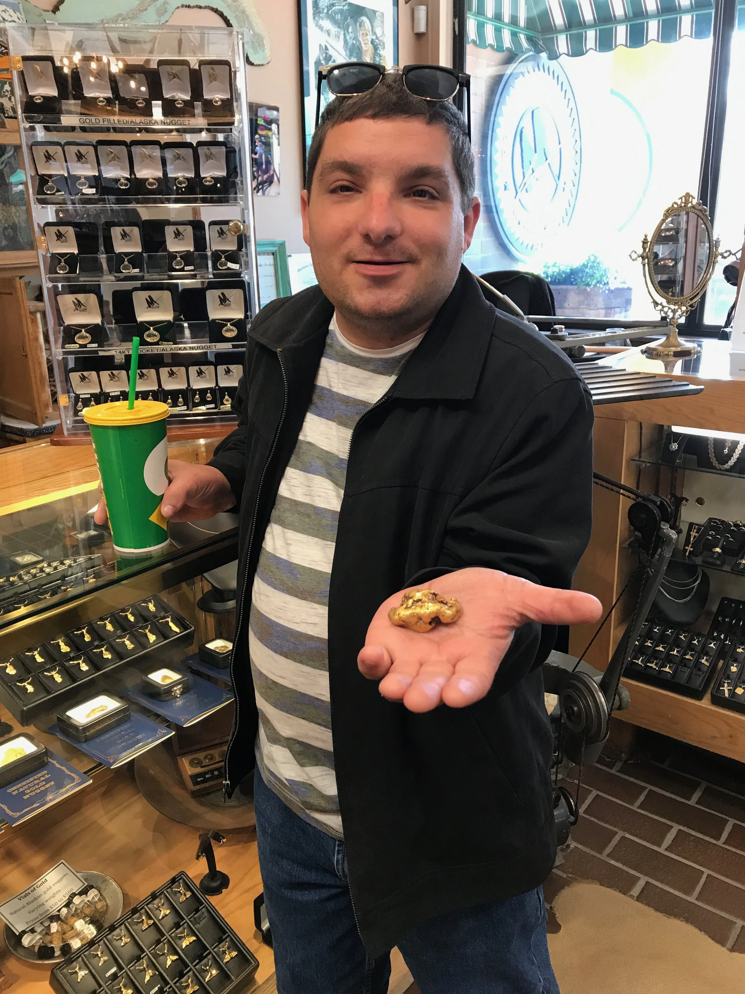 Michael holding a large gold nugget in his hand at a jewelry store. He has sunglasses on his head and is holding a green drink with a yellow lid and green straw in his other hand. The background displays jewelry items and a display case.