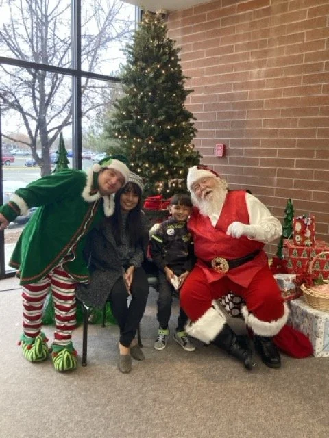 Michael dressed as an elf taking a photo with Santa Claus in front of a decorated Christmas tree at a holiday event.