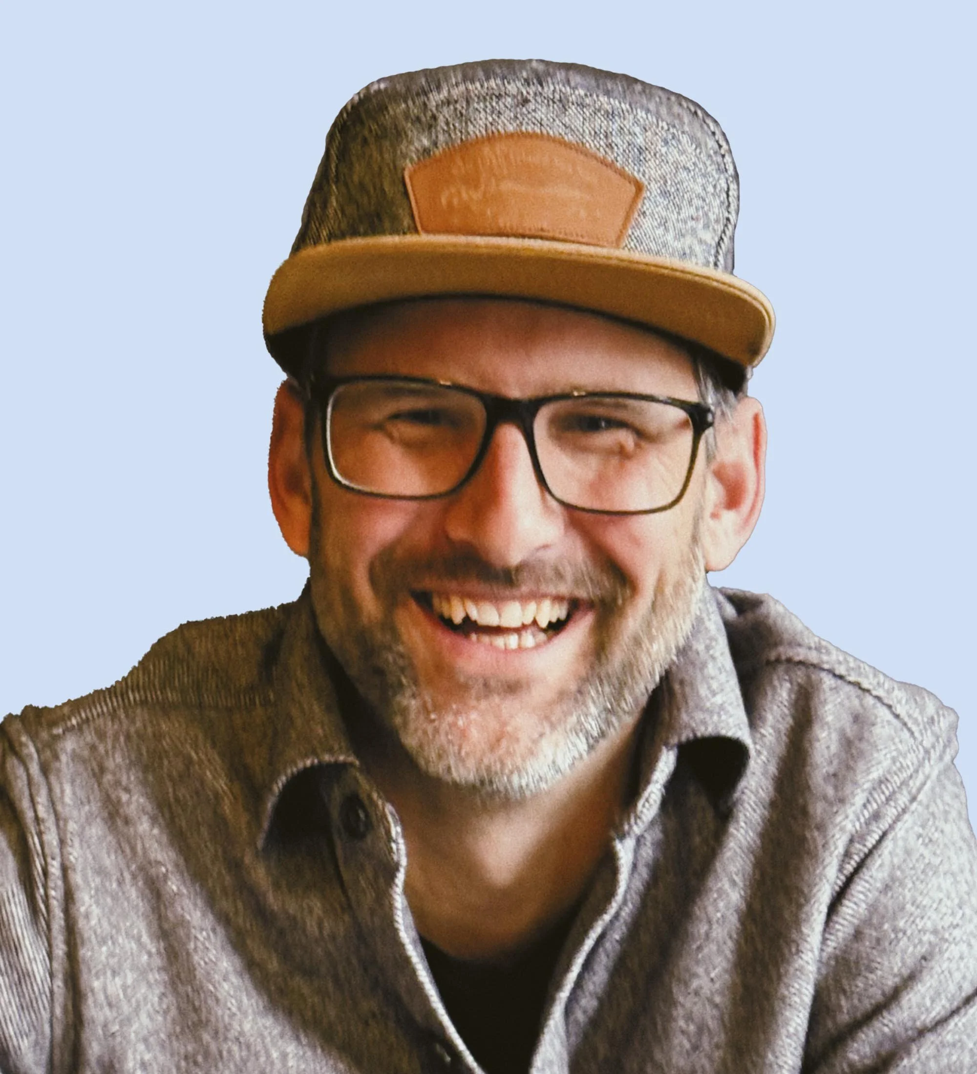 Board member Nate. A smiling man wearing glasses, a gray shirt, and a tan baseball cap with a leather patch, against a light blue background.