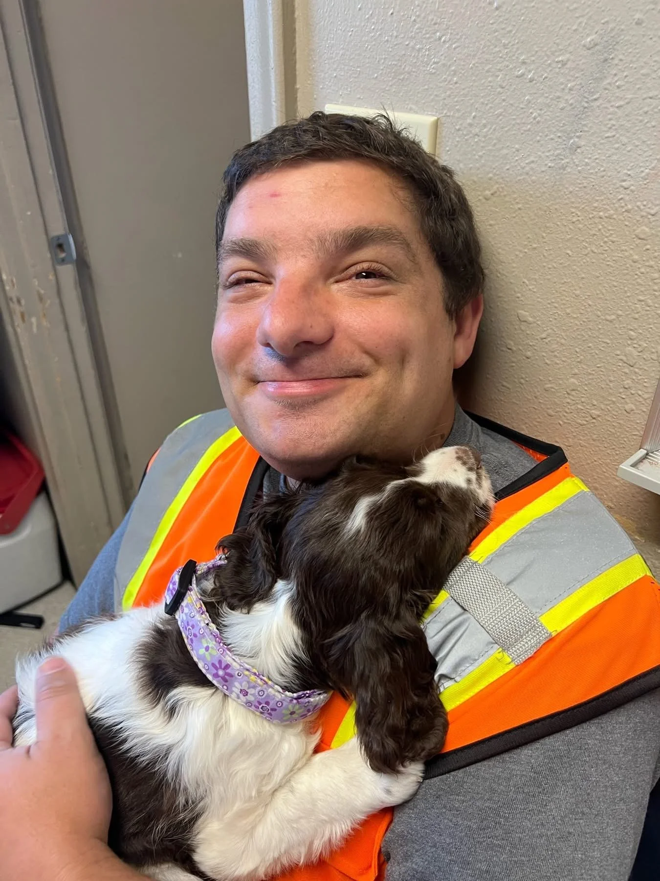 Michael smiling, wearing a gray shirt and a high-visibility safety vest, holding a small black and white puppy with a purple collar, resting its head on his shoulder.