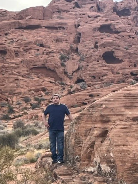 Michael standing on a red rock formations in a desert landscape with layered cliffs in the background.