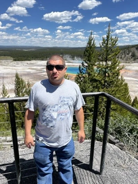 Michael wearing sunglasses and a gray t-shirt standing on a viewing platform with railing, overlooking a geothermal area with hot springs, steam, and colorful mineral deposits, surrounded by trees and blue sky with clouds.