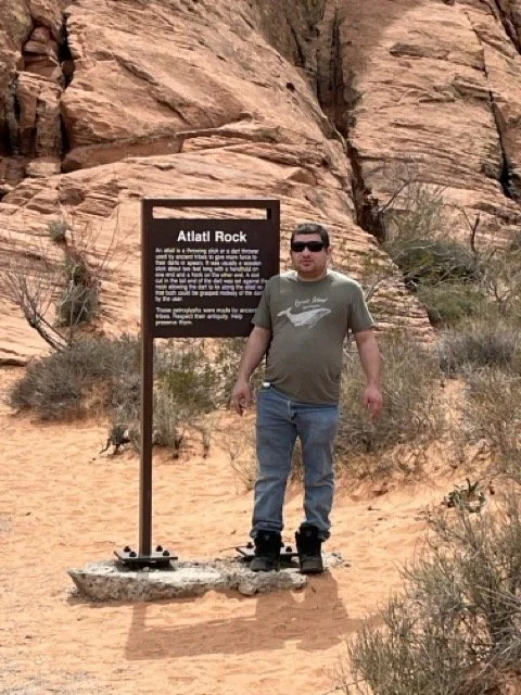 Michael standing next to an informational sign in a desert landscape with red sandstone formations and sparse vegetation.
