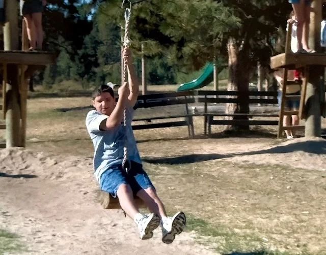 Michael as a boy swinging on a playground swing in a park with trees and a slide in the background.