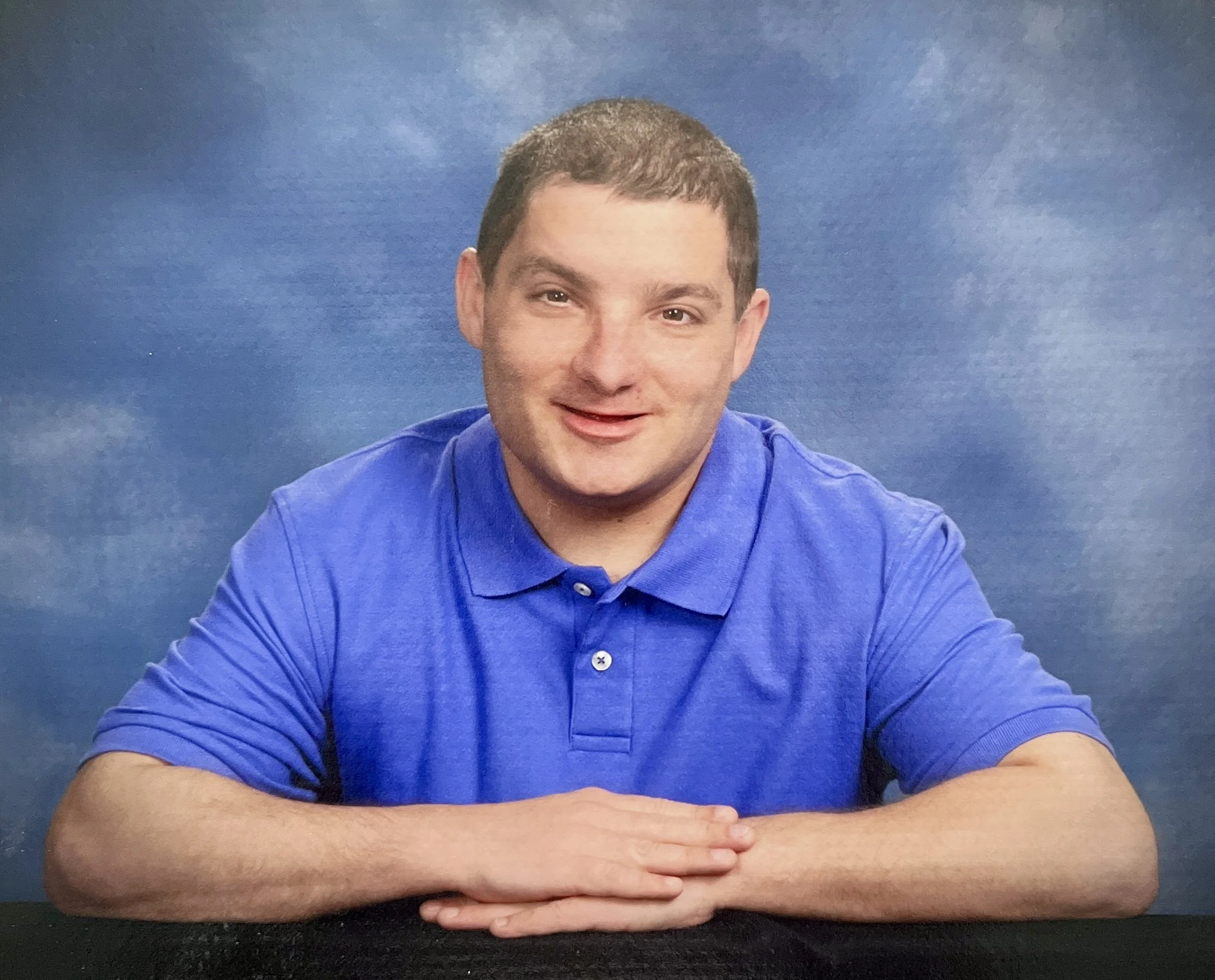 Michael as a young man with short brown hair, wearing a blue polo shirt, leaning on a surface with his hands crossed, smiling at the camera against a blue cloudy background.