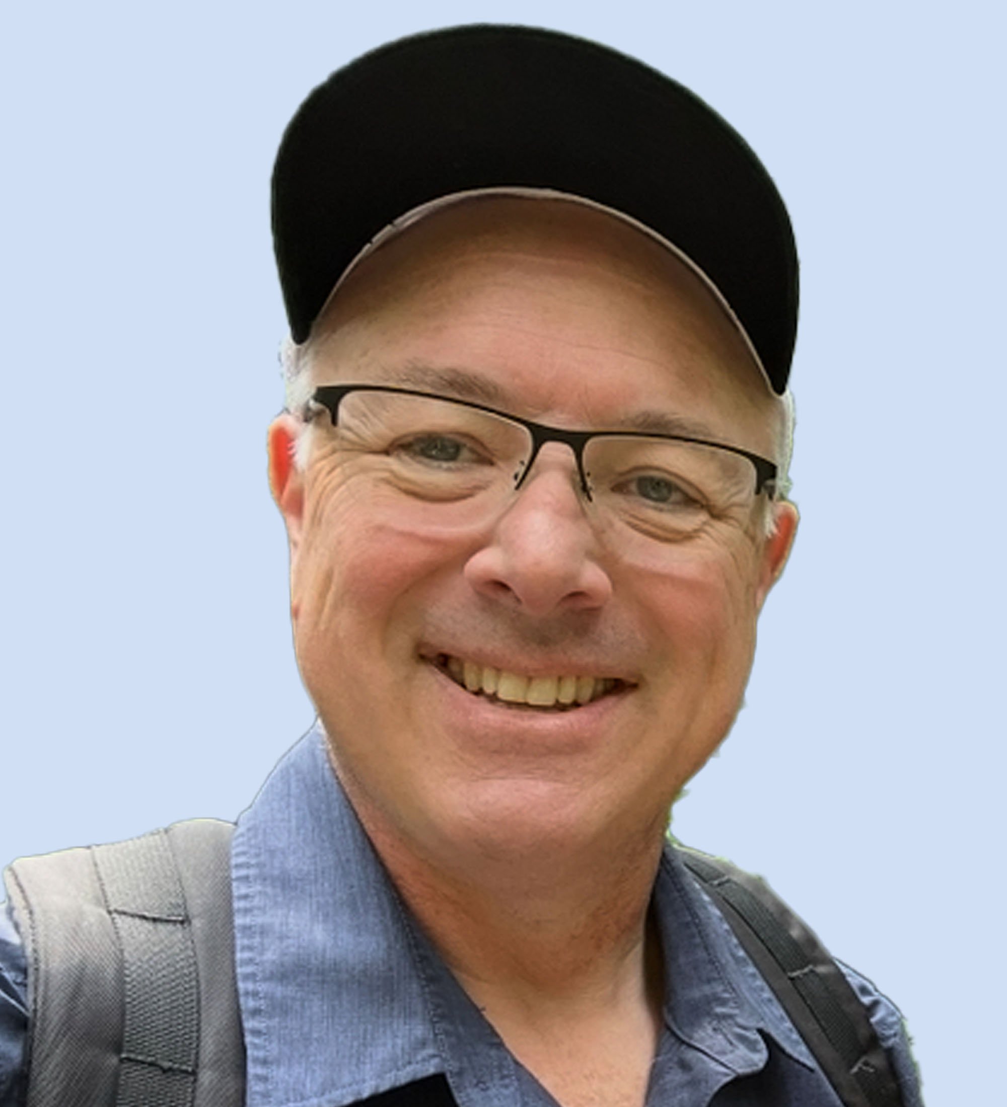 Board member Bruce. A smiling man wearing glasses and a black cap, with a backpack, outdoors against a clear light blue sky.