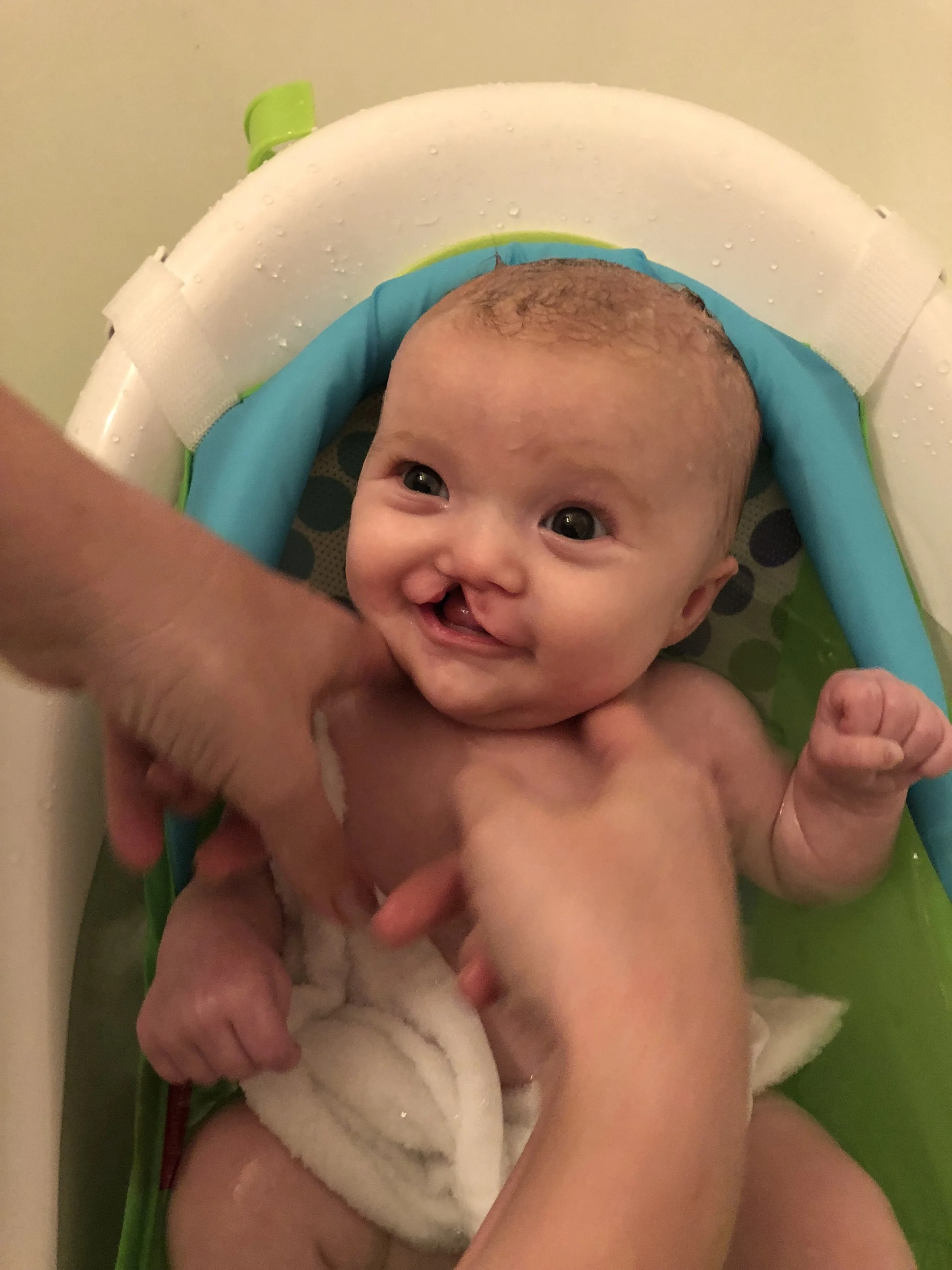 A smiling baby with wet hair and eyes wide open, lying in a baby bath with a white and green seat, being gently washed by an adult.