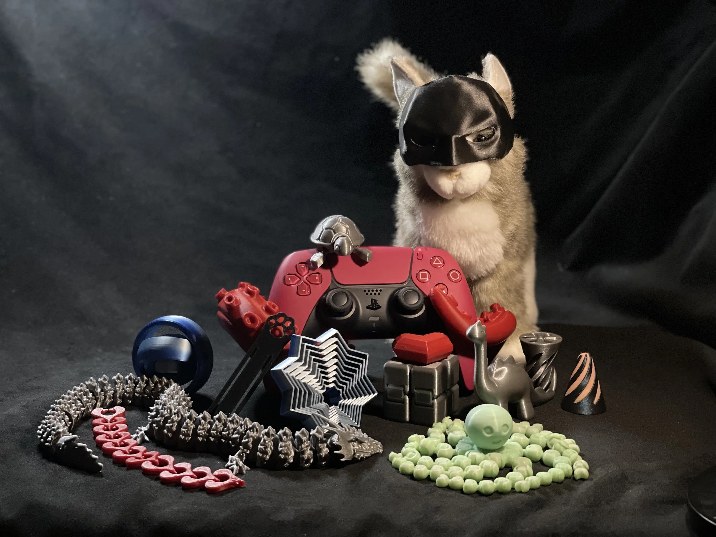 A cat wearing a Batman mask sitting behind an assortment of toys and figures, including a red PlayStation 5 controller, small figures, and decorative items, on a black surface with a black background.