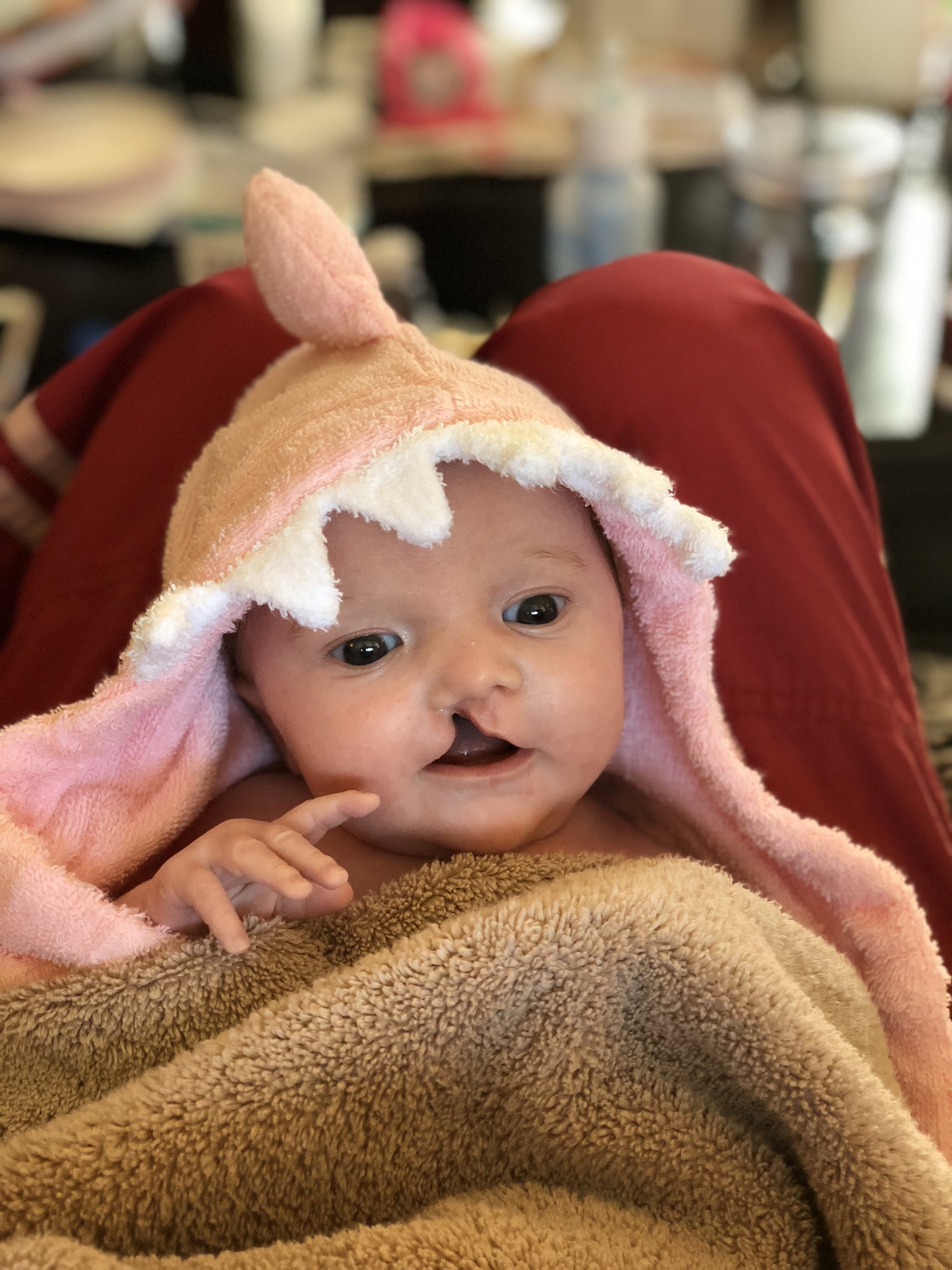 A baby wrapped in a brown towel with a pink hood that has a unicorn design, with a small horn and a mane, inside a room with blurred background.