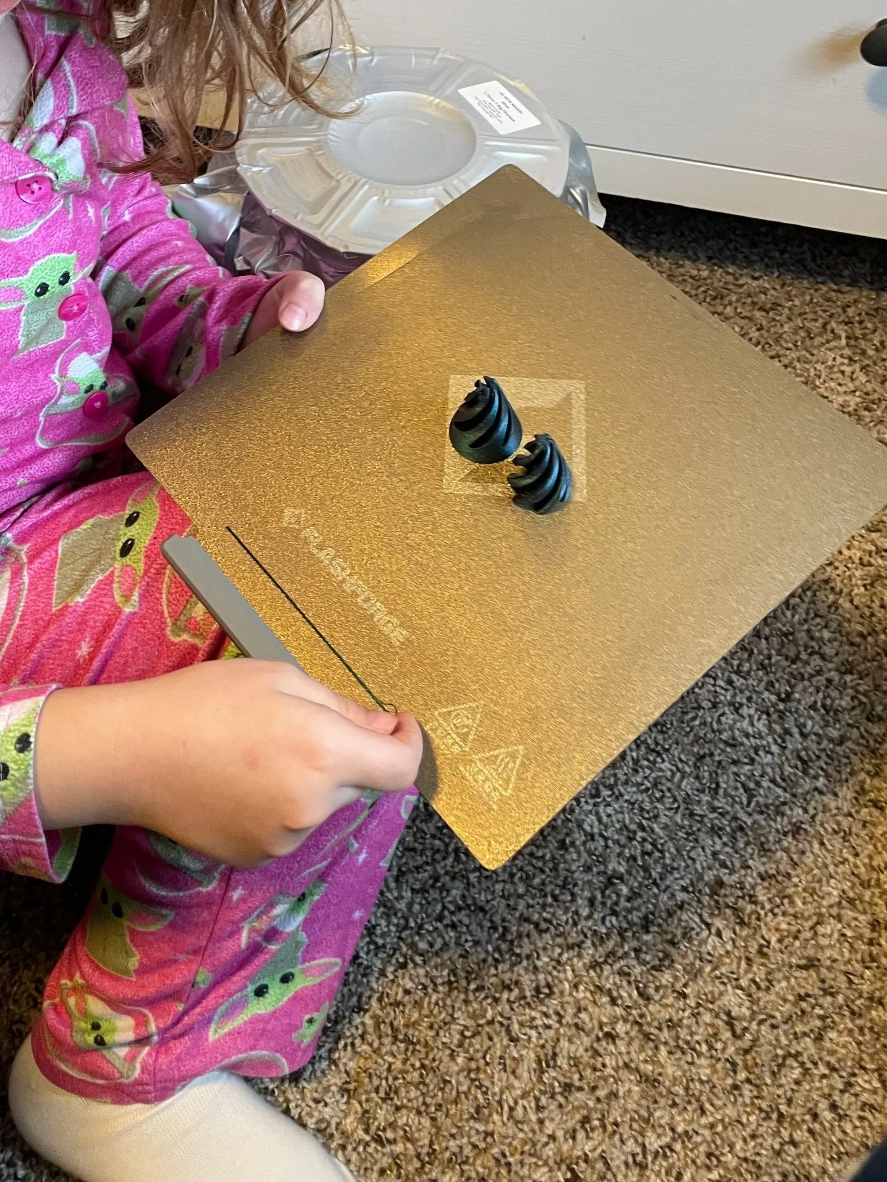 Child in pink pajamas with a frog pattern holding a gold-colored box with black, striped swirled object decorations on top.