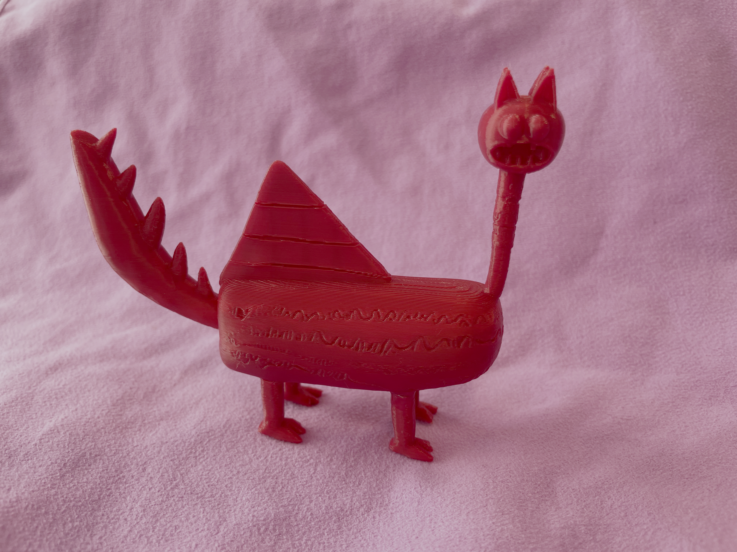 Red toy sculpture resembling a dragon with a triangular back and a long neck with a head that has cat-like ears and an open mouth, placed on a pink fabric background.
