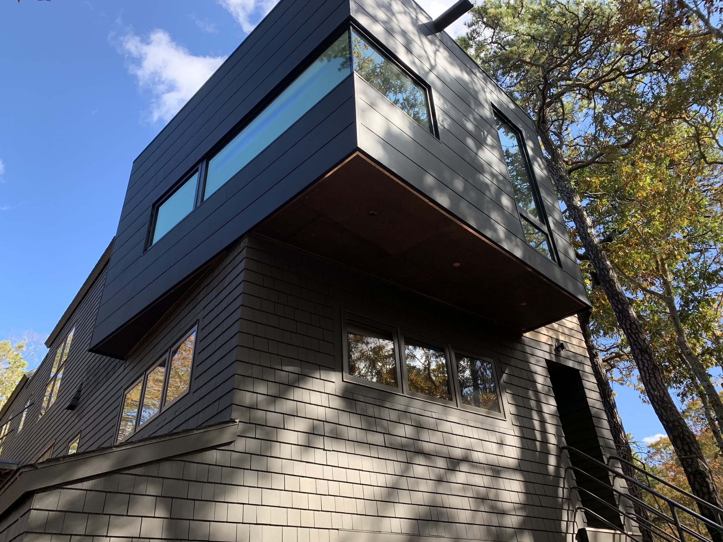 Modern black multi-story house with large windows, surrounded by trees with autumn leaves, under a partly cloudy sky.