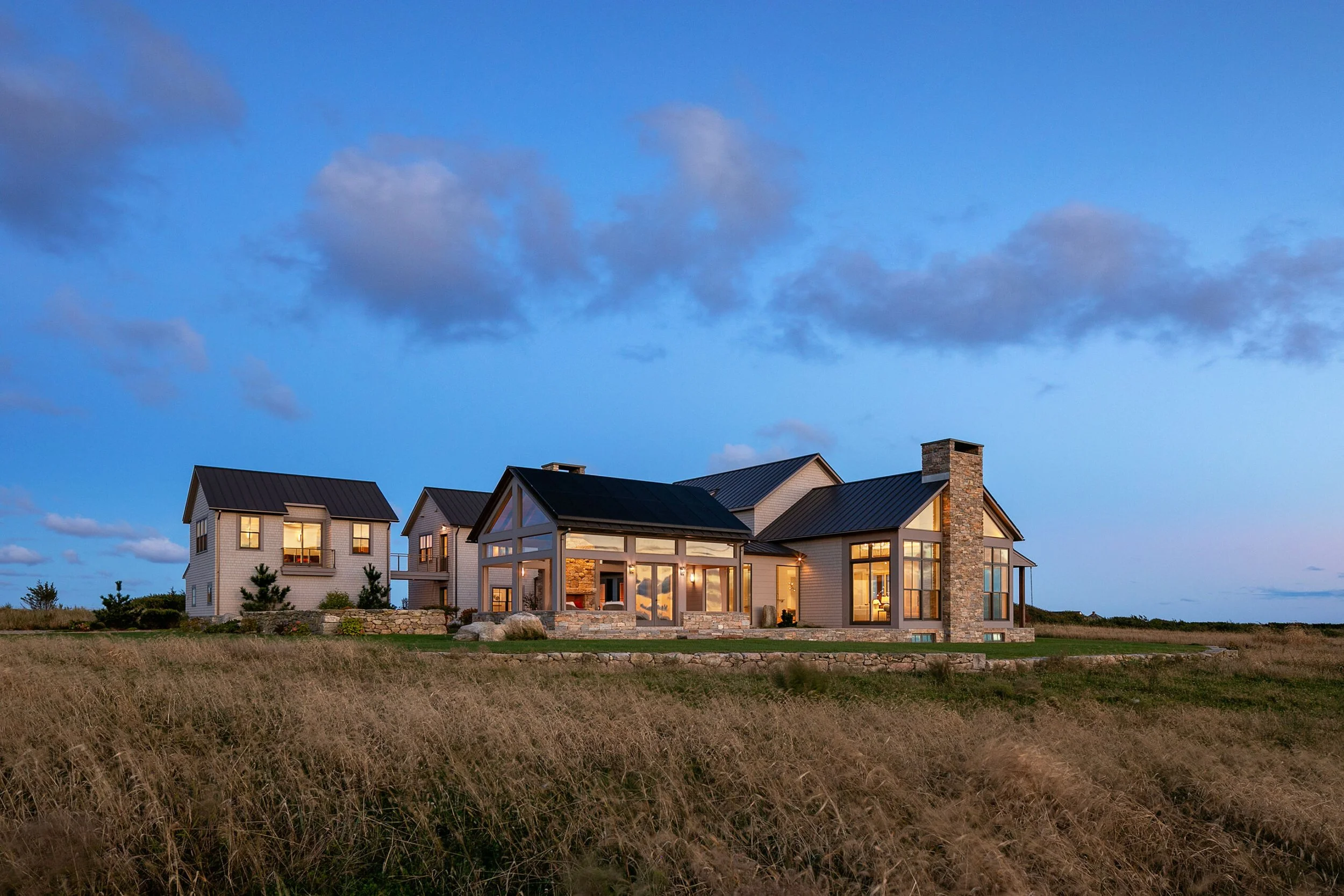 A large modern house with a combination of glass and stone exterior, situated in an open field during twilight with illuminated interior lights and a partly cloudy sky.