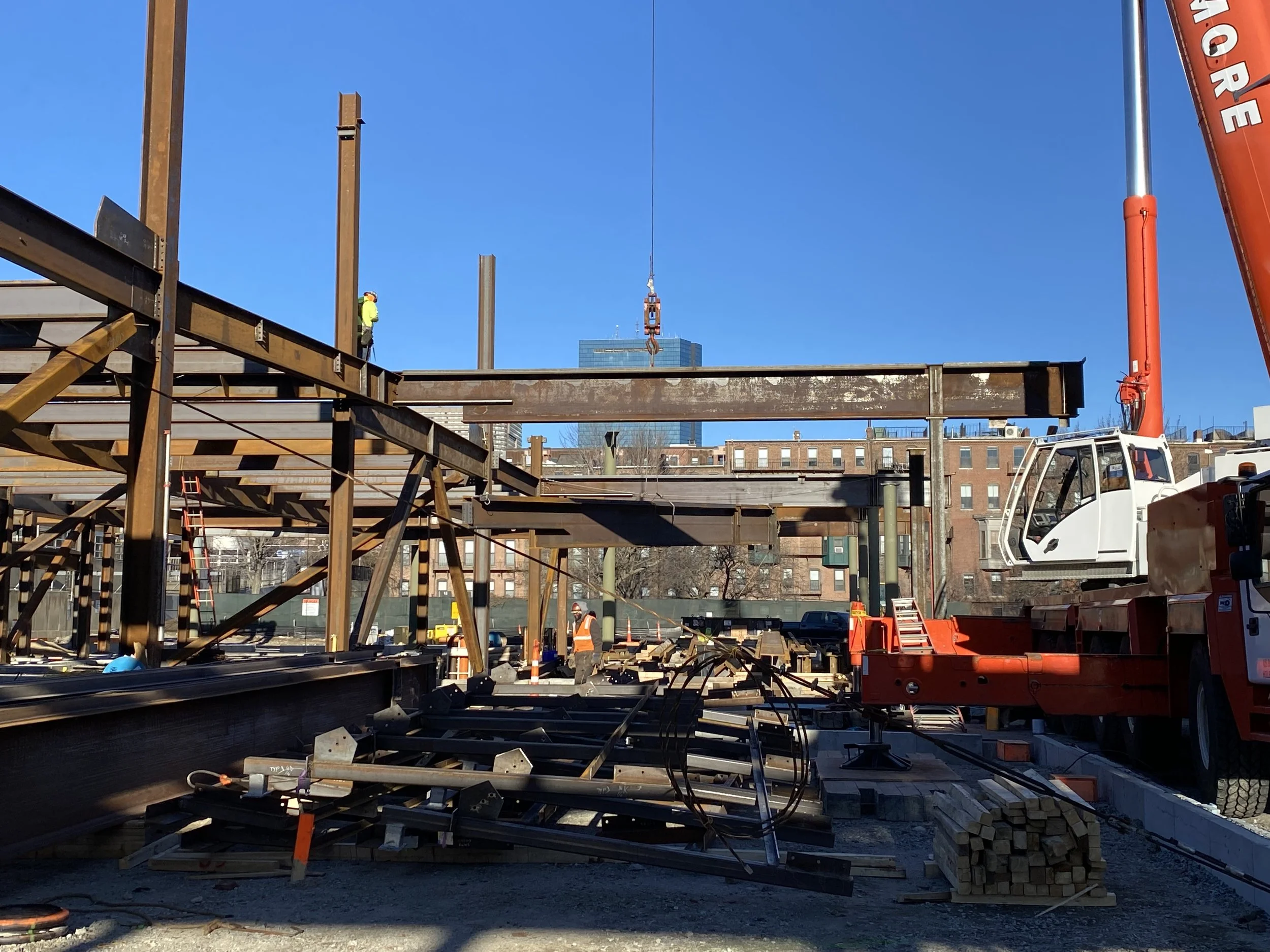 Construction site with steel beams, workers, and a crane lifting a steel beam in a city setting.