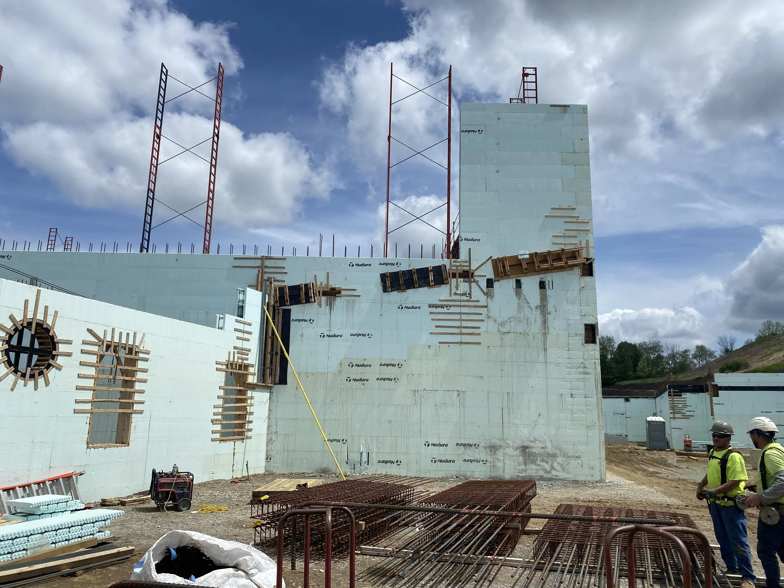 Construction site with workers wearing safety helmets and vests, a partially built concrete structure, scaffolding, construction materials, and a cloudy sky.