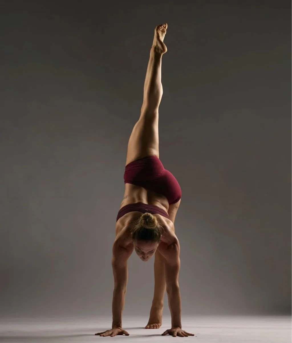 A woman performing a handstand with one leg extended vertically upwards, wearing maroon shorts and a sports bra on a plain background.